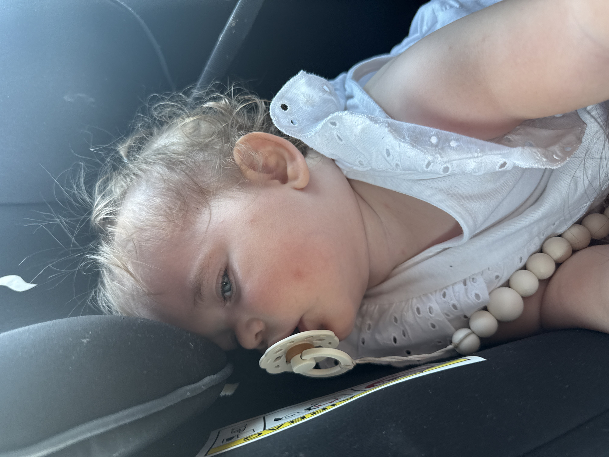 The image depicts a close-up view of a baby girl sleeping in a car seat. The baby has light-colored curly hair and is wearing a white eyelet-trimmed dress. A pacifier is in her mouth, and a string of wooden beads is visible around her neck. The car seat appears to be dark gray. A yellow and black warning label is attached to the car seat. The background is dark and out of focus, suggesting the interior of a vehicle. The image was taken in Lang-Göns, Germany.