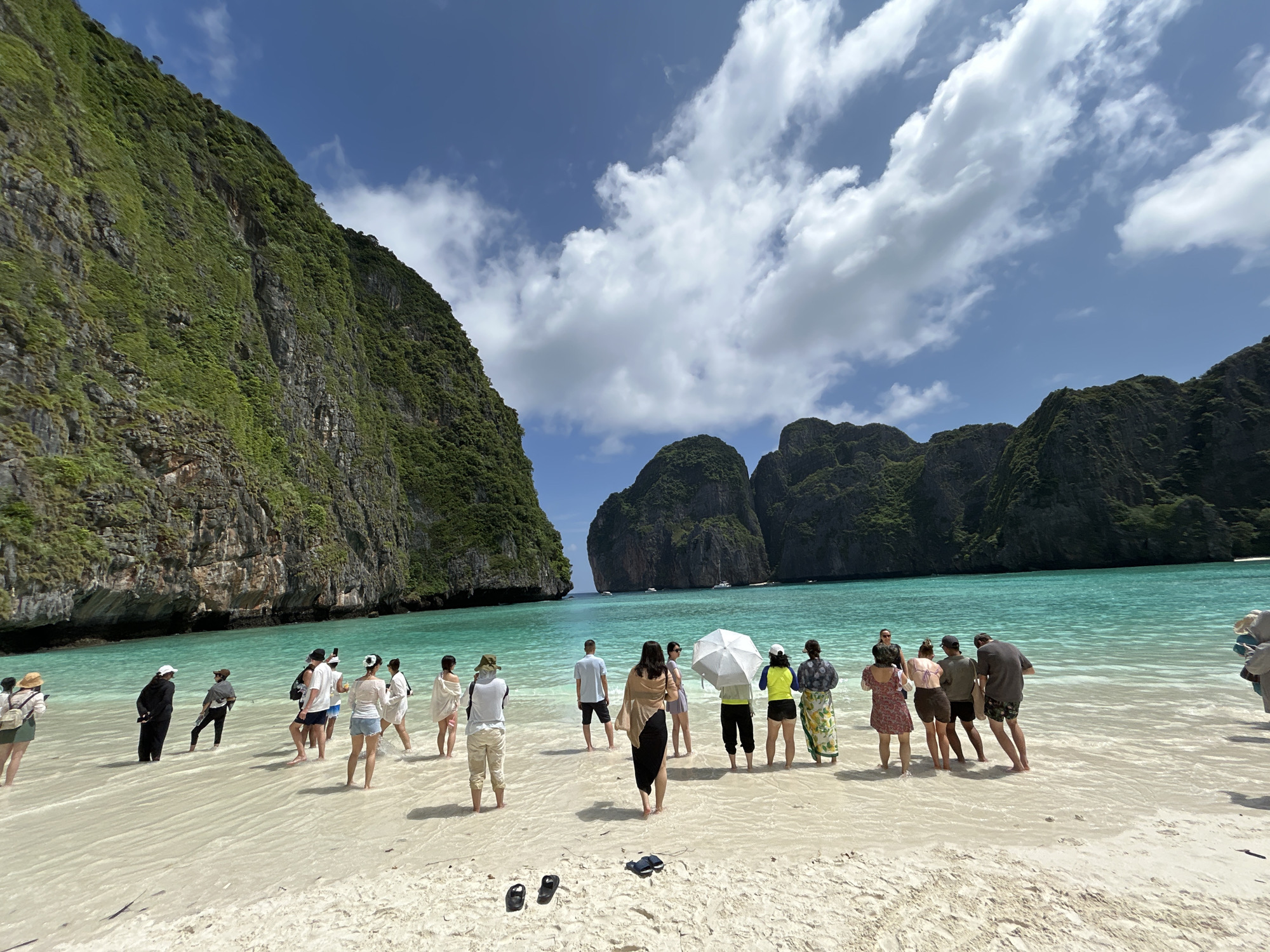 In Ao Nang, Thailand, a group of approximately 15 people are gathered on a sandy beach facing turquoise water. The beach is backed by steep, green, rocky cliffs. The sky is blue with scattered white clouds. The people are dressed in casual beach attire, with some wearing hats and one person holding a white umbrella. They appear to be a tour group, with most facing the water or the cliffs, seemingly taking in the scenery or posing for photos. Some individuals are wading in the shallow water. The sand is light colored and appears wet in places, indicating the tide is close to the shore. In the distance, the water is clear and the seabed is visible. Several small boats are anchored further out in the bay.