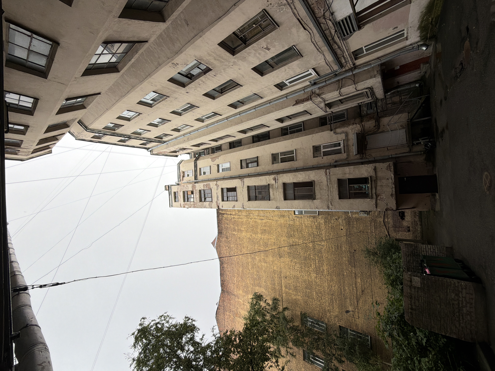 The image shows an interior courtyard in Saint Petersburg, Russia, with tall, narrow buildings surrounding it. The buildings are old, with weathered facades, and many windows. The sky is overcast. There is a large yellow brick building on the left, with a tree growing in front of it. In the center, there is a tall, thin building with many windows. On the right, there is another tall building with a more dilapidated facade. There are pipes and wires running along the buildings. In the foreground, there is a dark, paved courtyard with some puddles. There are also some trash bins visible. The scene is typical of the courtyards found in the historic center of Saint Petersburg.