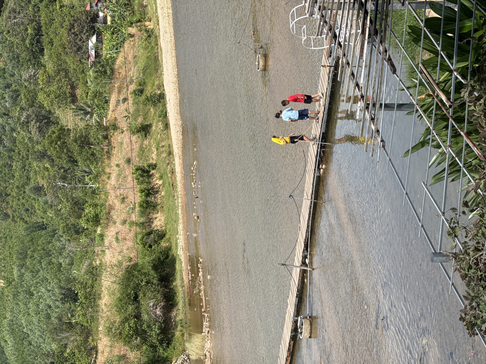 The image depicts a serene riverside scene in Nam Khanh Vinh, Vietnam. A rustic wooden boardwalk spans across the calm, clear water of a river. Three young individuals, two boys and a girl wearing a yellow headscarf, are walking on the boardwalk. The girl, in a yellow hoodie and black shorts, is positioned at the front left of the group. Behind her, a boy in a blue t-shirt and dark shorts is in the middle, and a boy in a red t-shirt and black shorts is on the right.
The riverbank is lined with coarse gravel and small rocks on the right side, and the left bank features lush green vegetation and trees. In the background, a verdant, tree-covered hillside slopes upwards, dotted with hints of distant buildings, suggesting a rural or semi-rural setting. The sky above is bright and clear, casting dappled sunlight onto the water.
In the foreground, part of a metal structure with horizontal bars and some greenery is visible, suggesting a viewing platform or a shaded area. Several circular concrete structures are partially submerged in the river, likely part of a farming or aquaculture setup. A white, arched structure, possibly a part of a water wheel or aeration system, is situated on the right side of the boardwalk. The overall impression is one of natural beauty and peaceful activity.