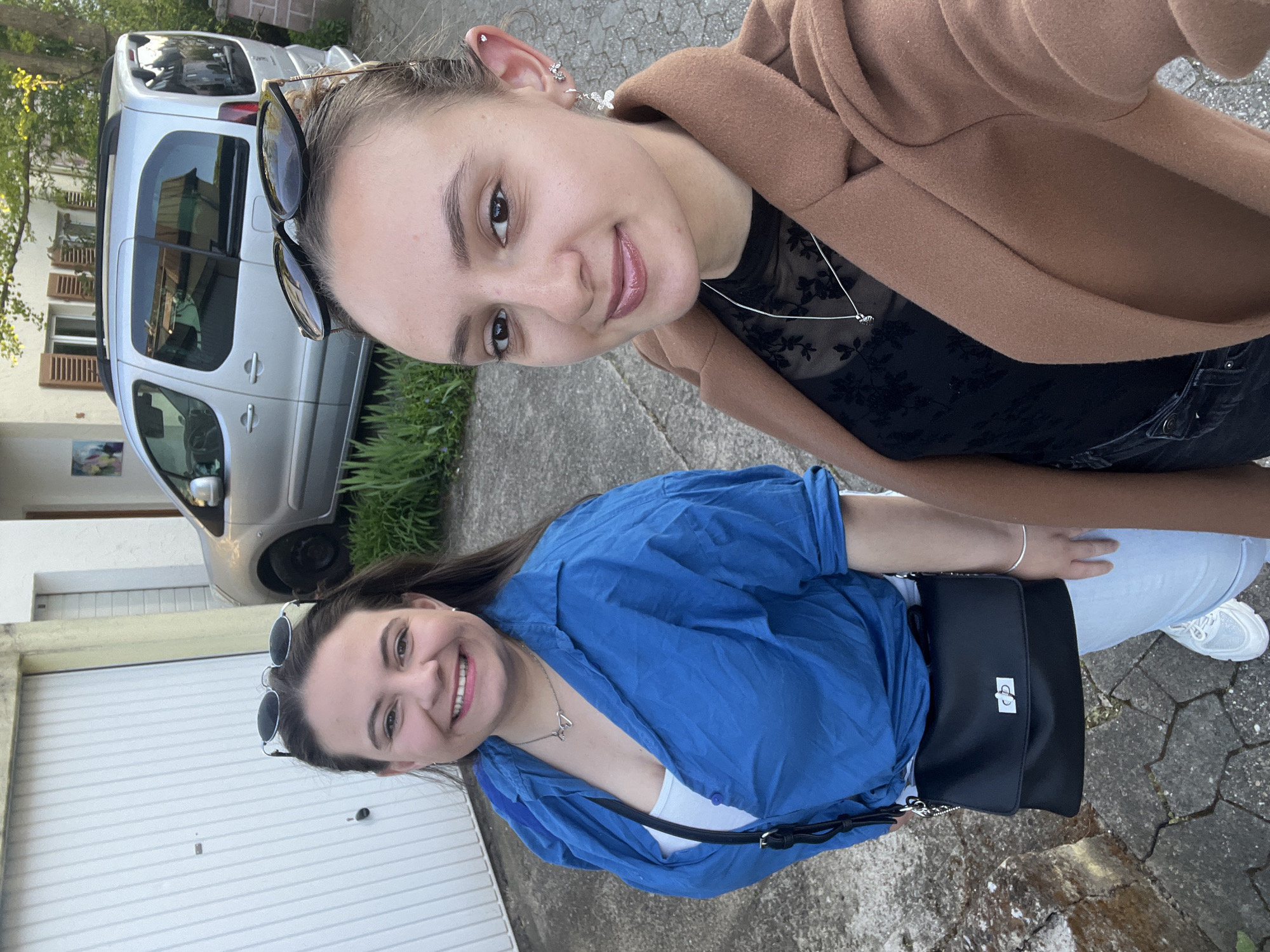 This daytime image, taken in a residential area of Saarbrücken, Germany, shows two young women posing outdoors. The photo is rotated 90 degrees clockwise, presenting the subjects and background sideways.
The woman positioned on the right (top when viewed in standard portrait orientation) has dark hair tied back, with black sunglasses resting on her head. She is smiling, revealing light makeup and glossy lips. Her ear features multiple piercings, including a stud and a delicate butterfly dangle earring. She wears a light brown coat over a black mesh top with floral embroidery, and a thin silver necklace with a small pendant.
The woman on the left (bottom when viewed in standard portrait orientation) also smiles warmly at the camera, with her dark hair loose and sunglasses on her head. She is dressed in a vibrant blue, long-sleeved button-up shirt worn open over a white top. A silver necklace with a "Love" pendant is visible around her neck, and she wears a silver bracelet on her wrist. Her lower attire consists of light-colored jeans and white sneakers. A black shoulder bag with a silver clasp is slung across her body.
The background reveals a typical residential setting. A silver car, resembling a Renault Kangoo-like van, is parked on a paved surface. Behind the car, a white house facade with closed brown window shutters and some green plants and trees can be seen. On the far left, a white garage door with horizontal paneling is prominent. The ground is paved, suggesting a driveway or street. Both women appear cheerful and relaxed, sharing a friendly moment.