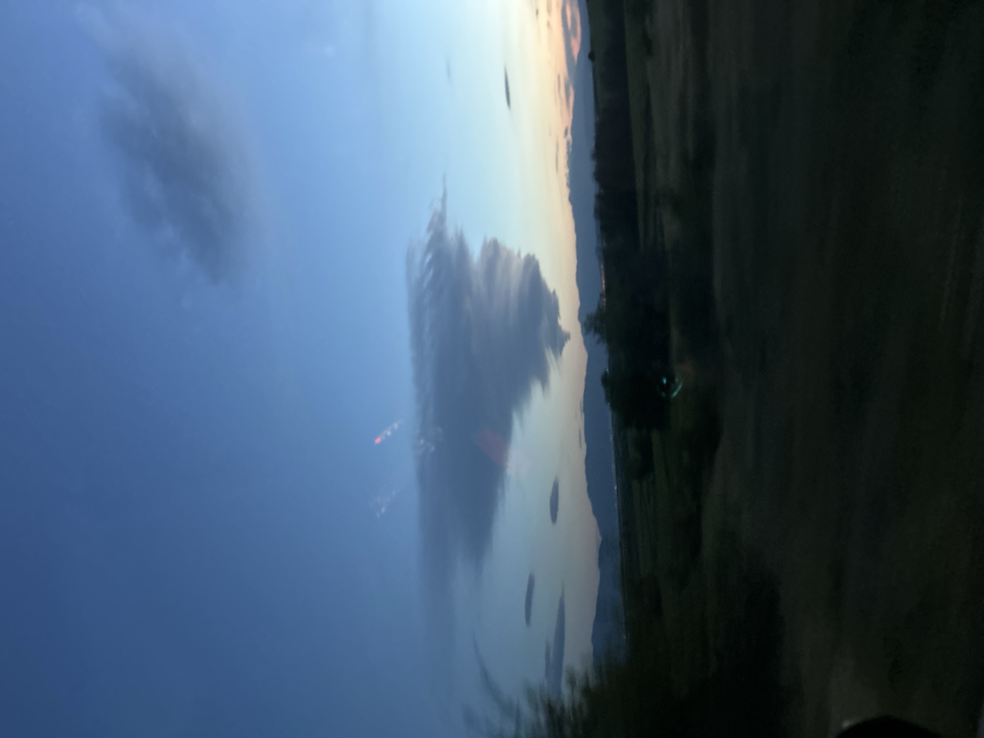 The image captures a landscape with a sky filled with clouds. The sky transitions from a bright blue on the left to a soft orange-yellow near the horizon on the right. There is a large, dark cloud formation in the center, with red lights that appear to be from city lights or traffic, also visible above the lake. A body of water reflects the sky and clouds. On the right, there is a dark green area that appears to be land or vegetation. The city of Ovchepoltsi is not immediately identifiable in this image.