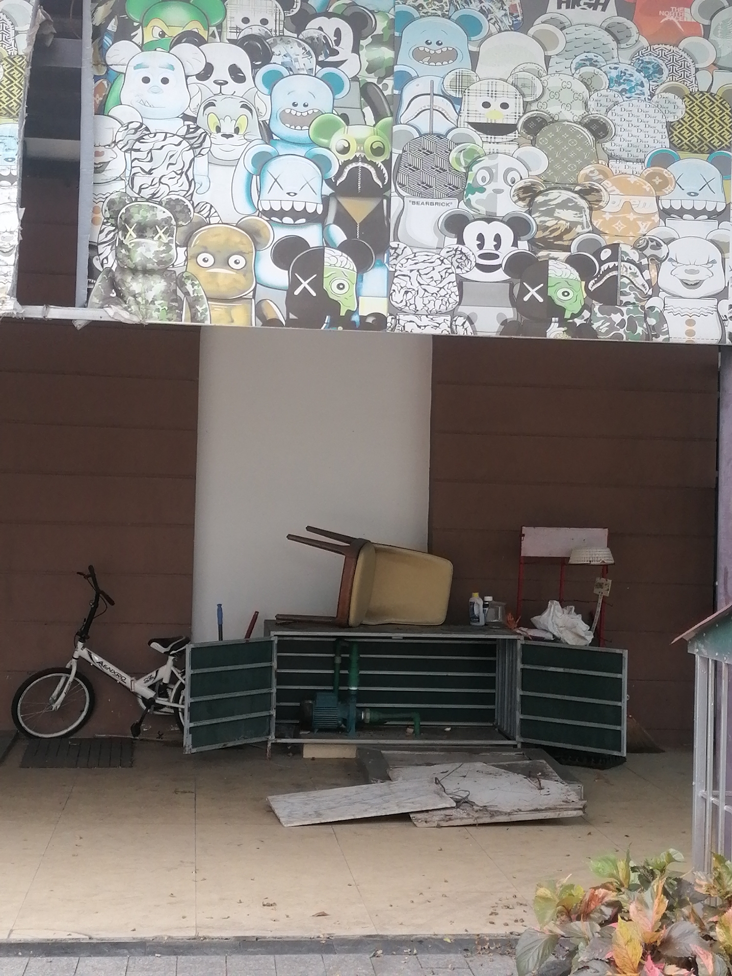 This full-shot image captures an outdoor area in Bai Chay Ward, Vietnam, featuring a vibrant, pop-art style mural above a utilitarian ground-level scene.
The upper portion of the frame is dominated by a busy mural or poster covering a building facade. It depicts numerous stylized bear-like figures, reminiscent of Bearbrick and Kaws art, each with unique patterns, colors, and often X-shaped eyes. Visible patterns include camouflage, stripes, polka dots, and iconic brand logos such as "Gucci," "Dior," "Louis Vuitton" (LV monogram), "Fendi," "BAPE," "The North Face," and "Nike" (swoosh). One figure clearly has "BEARBRICK" written on its chest, and another wears a cap with "HIGH." The figures are densely packed, creating a collage effect.
Below the mural, at ground level, the scene transitions to a more practical and somewhat disheveled space. A white bicycle with "AENXRO" printed on its frame leans against a wall with horizontal brown paneling on the left. In the center, an open metal cabinet or storage unit, dark green with horizontal slats, stands open. A light-colored chair is overturned on top of the cabinet. Inside the cabinet, a blue water pump is visible. Adjacent to the cabinet, a few bottles, a white cloth, a small white stand, and a broom are scattered. On the tiled ground in front of the cabinet, several pieces of broken, light-colored board or debris are present. The wall on the right side of the ground level is mostly white, while a small structure with a gabled roof and white framing is partially visible on the far right.
The overall lighting suggests it is daytime, possibly overcast, given the lack of strong shadows. No direct human activity is taking place, presenting a static scene of urban exterior and storage. Lush green and reddish foliage is visible in the bottom right corner, indicating outdoor surroundings.