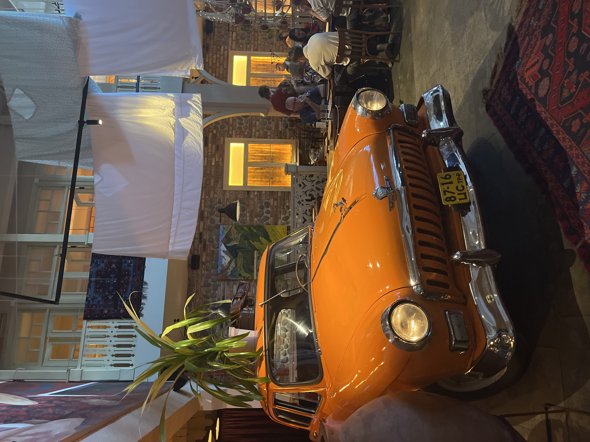 The image shows an indoor scene, possibly a restaurant or event space, featuring a vintage orange car as a prominent decorative element. The car is positioned in the foreground, facing the viewer, with a plant growing from its roof. In the background, several people are seated at tables, engaged in dining and conversation. The environment appears to be decorated with a rustic aesthetic, incorporating exposed brick walls, wooden furniture, and draped fabrics. A Persian rug is visible on the floor in front of the car. The lighting is warm and ambient, suggesting an evening setting. The location is in Unknown, Unknown.