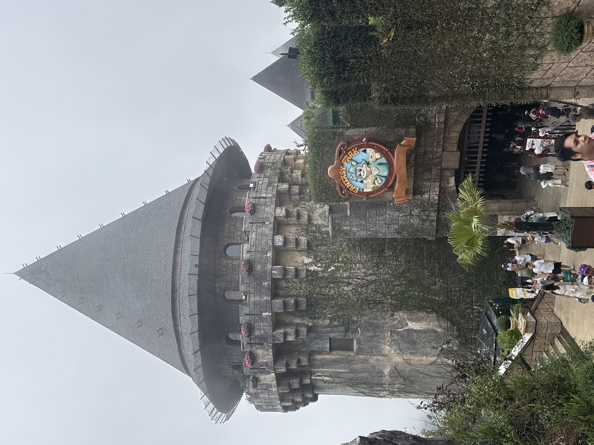 The image captures a view of Sun World Ba Na Hills in Đà Nẵng, Vietnam. The foreground is dominated by a medieval-style stone tower with a conical roof, typical of the park's architecture. The tower features small windows, decorative flowers, and climbing vines. To the right of the tower is a stone structure with an archway leading into what appears to be a restaurant or dining area. Above the archway is a sign reading "Fantasy Park," decorated with cartoonish figures. People are seen entering and exiting through the archway. The background shows more of the mountainous landscape and other architectural structures. The sky is overcast, suggesting cloudy weather. The setting is a theme park with fantasy-themed architecture and tourist attractions.