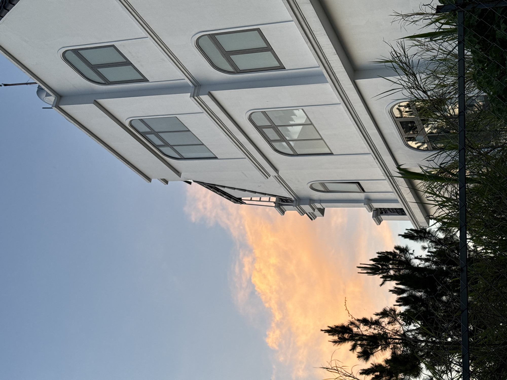 The image, taken in Lâm Viên - Đà Lạt, Vietnam, shows a low-angle perspective of a building with a white exterior. The building has multiple floors, with distinct windows featuring arched tops and divided panes. The sky, transitioning from blue to orange hues suggestive of either sunrise or sunset, serves as the backdrop to the building. Silhouetted trees and some greenery are partially visible on the right side of the frame, behind a chain-link fence.