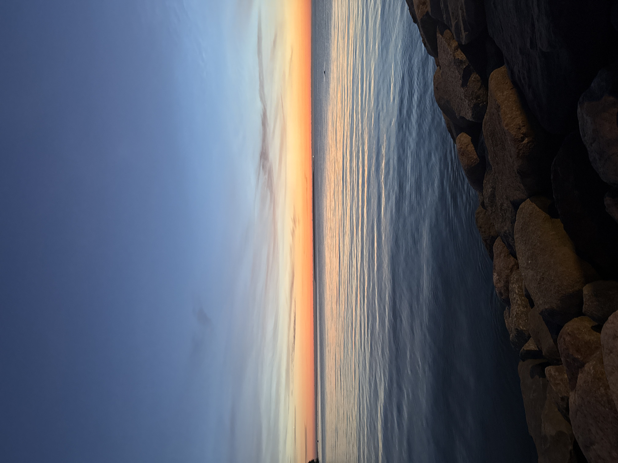 This is a photograph of a sunset over the sea, with a rocky shoreline in the foreground. The sky transitions from a deep blue at the top to hues of orange and yellow near the horizon, indicating the sun has recently set or is about to set. Wispy clouds are scattered across the sky. The sea is calm with gentle ripples reflecting the colors of the sky. Large, rough rocks form a barrier along the right and bottom edges of the image, partially illuminated by the fading light. In the distance, along the horizon, faint landmasses can be seen. A small, dark silhouette, possibly a boat or a bird, is visible on the water. The scene appears tranquil. This image is not identifiable as being located in Tallinn, Estonia.
