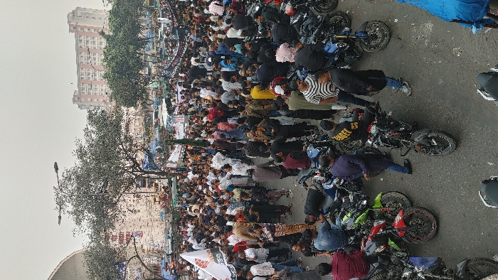 A large crowd of people stands shoulder to shoulder alongside parked motorcycles on a city street. Buildings rise in the background. Some individuals wear light-colored clothing. The scene appears to be a gathering or protest. A tall streetlight fixture is visible amidst bare trees. The sky is overcast. A large banner is held up by some of the people in the crowd.