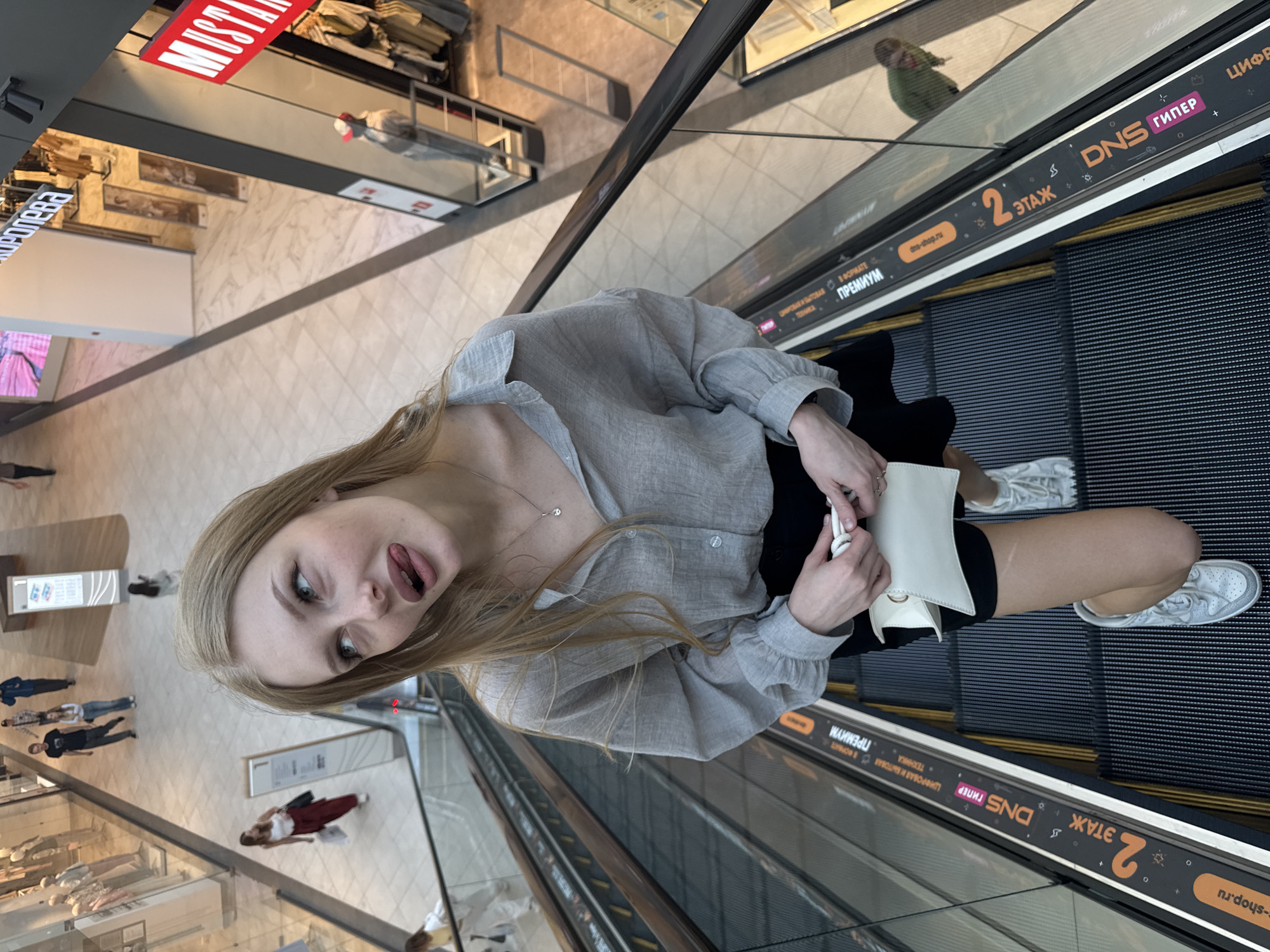 The image shows a young woman on an escalator in a shopping mall. She has blonde hair and is wearing a gray shirt and a black skirt. She is holding a white clutch bag and sticking her tongue out at the camera. The escalator has advertisements on the side for "DNS" and "2 ETAGE," which are brands and a floor number in Russian. The shopping mall appears to be modern with marble floors and shops visible in the background. The woman's pose and expression suggest a playful or humorous mood. It's impossible to definitively determine the city as Perm, Russia, solely from the visual information provided in the image.