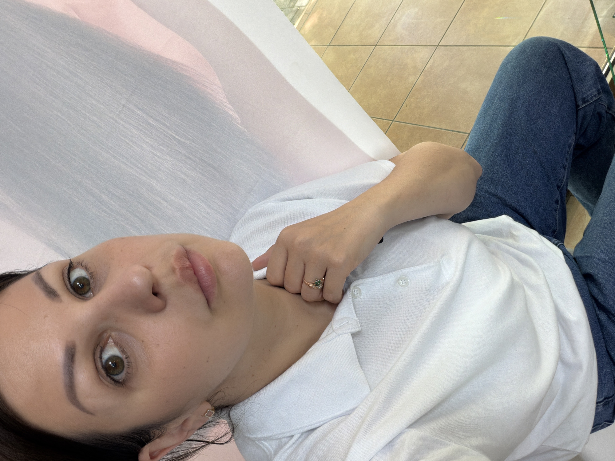 A woman with dark hair and brown eyes is taking a selfie in an indoor setting with tiled flooring. She is wearing a white polo shirt and blue jeans. She has a ring with a green stone on her right hand, which is touching her neck. The background is a light-colored wall with vertical lines and a pinkish hue. The lighting suggests an indoor space, possibly a commercial establishment, given the tiled floor and the backdrop. There is no indication of the city of Sochi, Russia.
