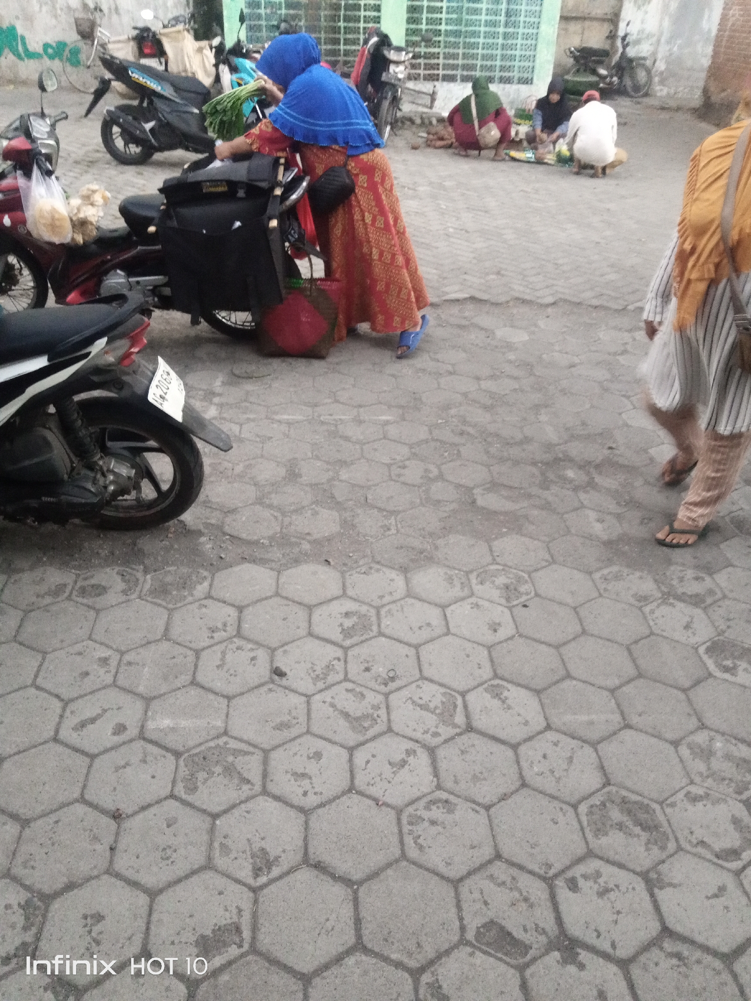 The image depicts an outdoor scene on a paved ground surface in what appears to be a market or roadside area. In the foreground, a portion of a white and black motorcycle is visible, featuring a license plate that reads "AG 2069". Two individuals are centrally positioned. One, wearing a blue hijab and a red and gold patterned dress, is leaning over a black bag attached to a motorcycle. Another person in a blue hijab is also present, reaching for something green, likely produce, from the motorcycle. Further in the background, three individuals are crouched, appearing to be vendors or shoppers, with various items laid out on the ground. One person is wearing a green head covering, another a black head covering, and a third a white top. The environment shows a mix of buildings and a wall with graffiti in the upper left corner. The ground is paved with hexagonal concrete blocks. The overall impression is of a bustling, everyday scene in a public space.