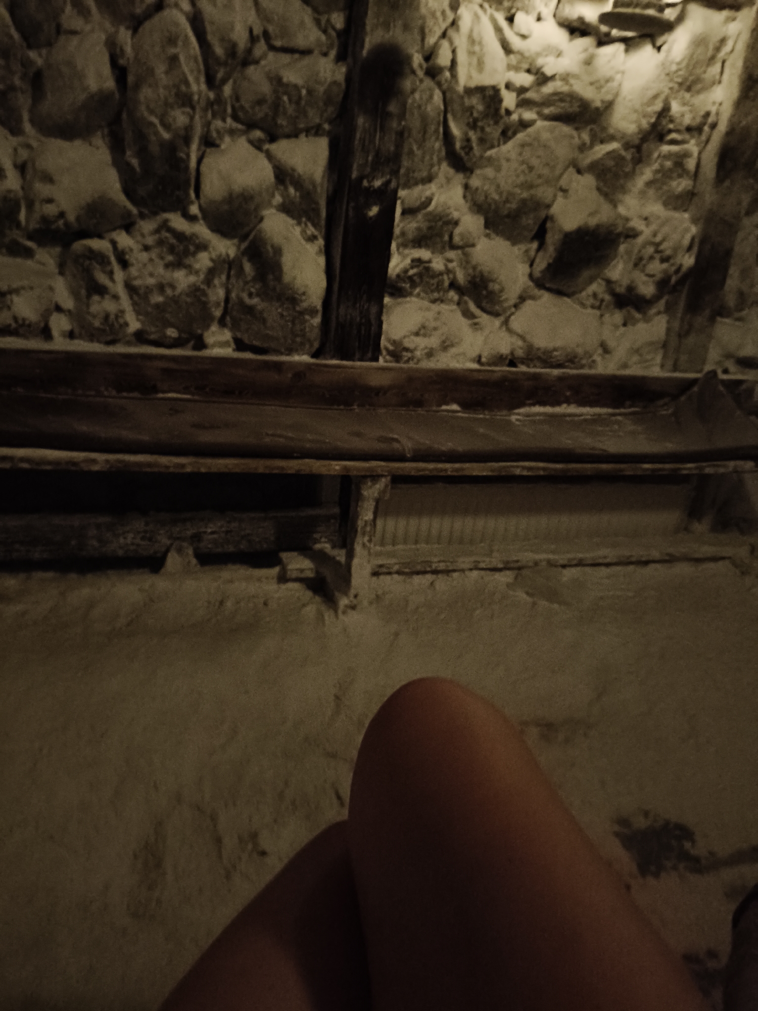 An indoor scene captured from a low angle, featuring a rough-hewn stone wall as the backdrop. A wooden bench is suspended horizontally in the mid-ground, with its surface appearing worn. Below the bench, a grated or slatted surface is visible. The foreground is occupied by the legs of an individual, suggesting they are seated or reclining. The overall lighting is dim, with a warm, sepia tone dominating the image, creating a somber or ancient ambiance. The location is Plovdiv, Bulgaria, specifically within the Kophala Salt Cave, a therapeutic salt chamber known for its unique geological formations and mineral-rich environment.
