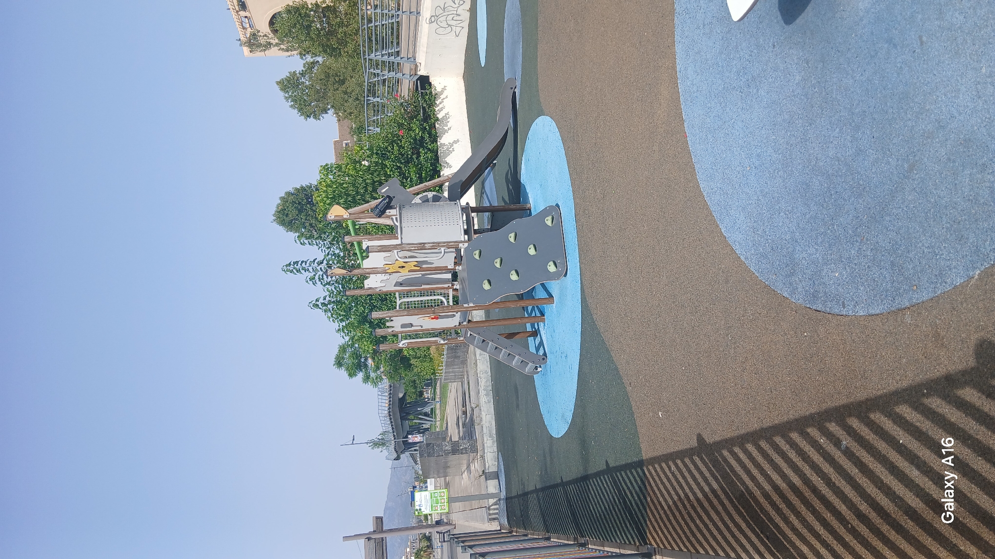 The image depicts a modern playground located in San Pedro de Alcántara, Spain. The playground features a multi-level play structure with a gray climbing wall, a slide, and stairs. The ground surface is a safety surface composed of blue and brown rubber material. In the background, a pedestrian bridge spans across a road, and various buildings and lush green trees are visible under a clear blue sky. A metal fence is present on the left side of the image, casting shadows on the ground. The overall scene suggests a public recreational area.