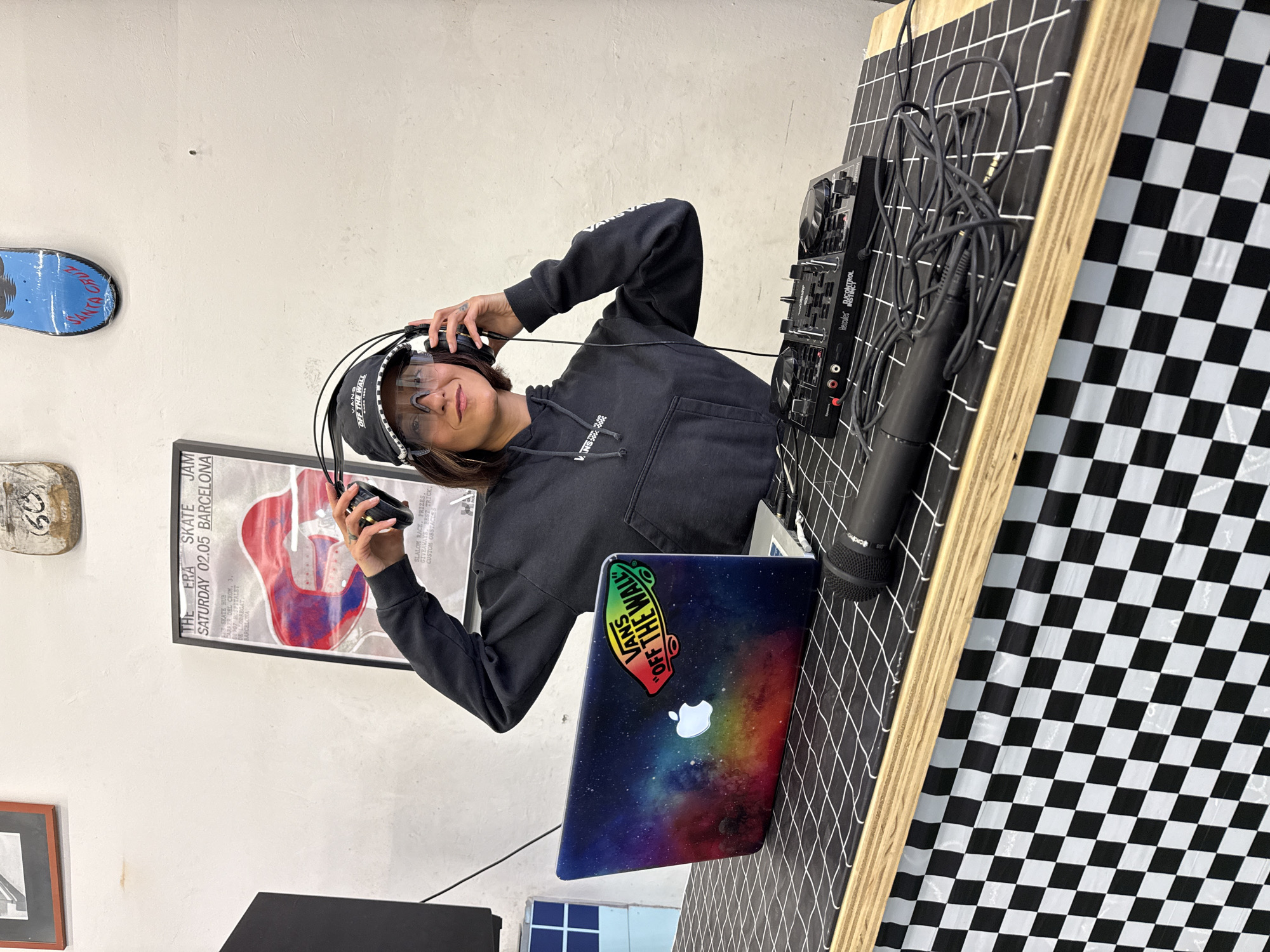 The image captures a young woman standing in front of a DJ setup, presumably in L'Hospitalet de Llobregat, Spain. She is wearing a black hoodie with the Vans logo, a Vans visor, and headphones, holding one side of the headphones up. The DJ setup consists of a mixer and a laptop with a colorful space-themed skin and a Vans sticker. A microphone also stands on the table. The DJ setup is placed on a wooden board covered with black and white checkered and grid-patterned material. The background includes a white wall with various skateboards and a poster with "THE ERA SKATE JAM SATURDAY 02.05 BARCELONA" printed on it.