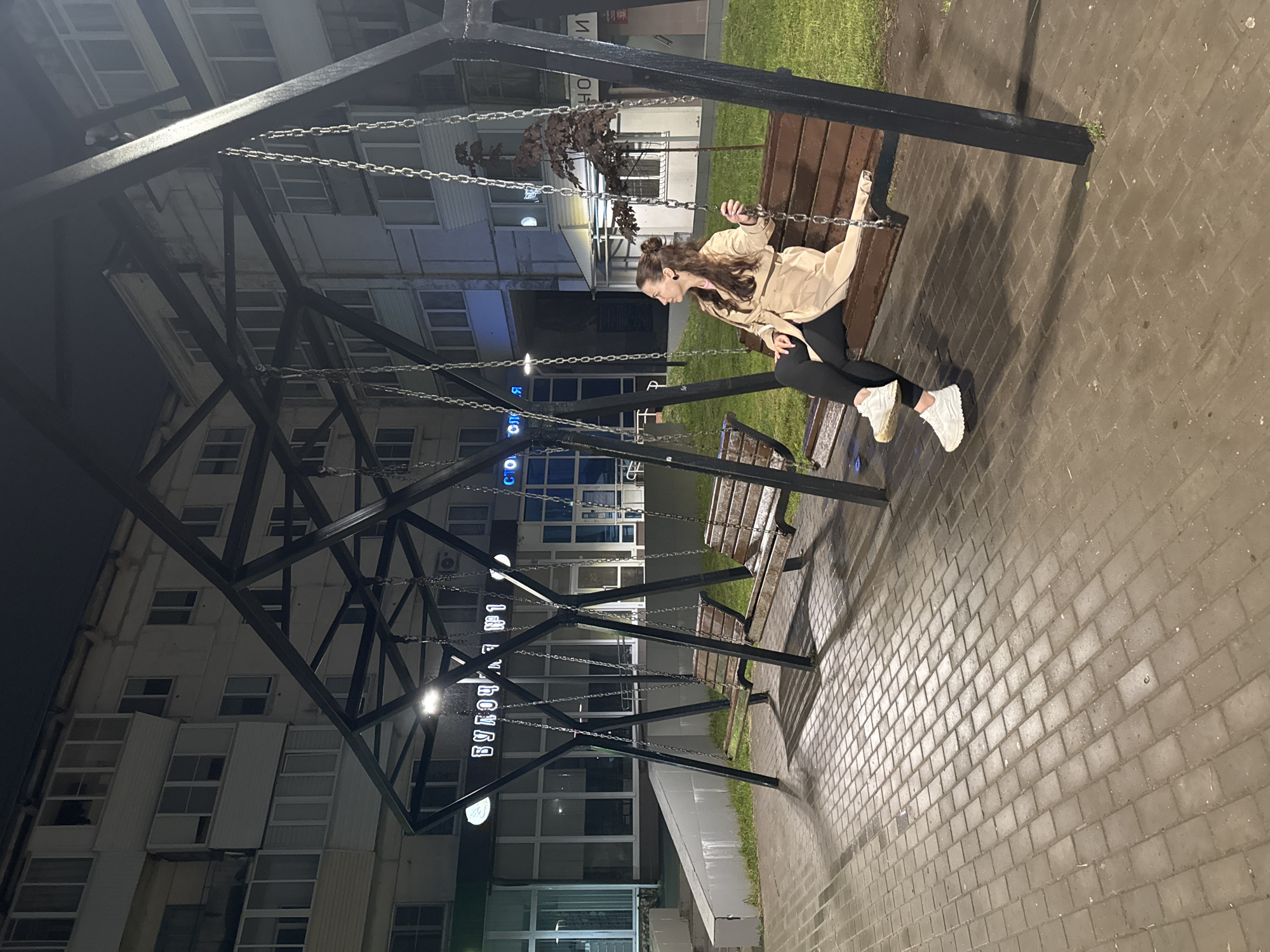 This image captures a nighttime urban scene in Chaikovsky, Russia. A single adult female, appearing to be in her late 20s or 30s, is seated in profile on the rightmost of three dark wooden swing benches. She has dark brown hair partially tied in a bun, wears a light-colored, long trench coat, black leggings, and white athletic shoes, and holds one of the swing chains with her right hand while gazing to the left.
The swing structure is a prominent, dark-colored metal installation with a geometric design, featuring interconnected beams that support the chain-hung benches. A bright LED light fixture is mounted centrally on the top of the swing structure, casting strong illumination onto the paving below and creating distinct shadows.
In the background, a multi-story, light-colored apartment building dominates the scene. Its facade shows signs of weathering, and it features numerous windows, some with enclosed balconies. The ground floor of this building houses commercial establishments, identifiable by illuminated signs with Cyrillic script: "БУЛОЧНАЯ № 1" (Bakery No. 1) is clearly visible, and a partial sign for "СТОМАТОЛОГИЯ" (Dentistry) is seen further to the right. A small, dark-foliaged tree or shrub is situated behind the swing structure on the right.
The ground area in the foreground is paved with rectangular grey interlocking pavers, which appear damp and reflective, suggesting recent precipitation or high humidity. Patches of green grass are visible bordering the paved sections. The overall scene is illuminated exclusively by artificial light sources from the swing structure and the background buildings, indicating that it is dark outside.