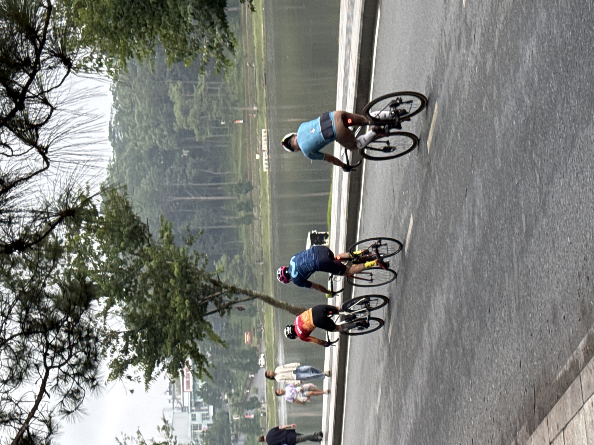 In Phường Xuân Hương - Đà Lạt, Vietnam, three individuals are cycling on a paved road beside a body of water. The cyclist in the foreground, wearing a blue jersey and brown shorts, is positioned facing away from the camera. To their left, two other cyclists are riding side-by-side. The cyclist on the right wears a red and yellow jersey, while the cyclist on the left wears a red helmet. In the background, two young women and one man are walking on a sidewalk adjacent to the road. Buildings are visible behind the walkers, and a densely wooded hillside overlooks the water. The sky is overcast.