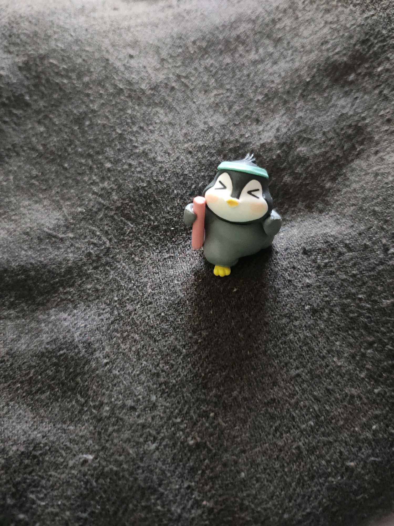 A close-up, high-angle shot shows a small, dark grey penguin figurine with yellow feet, a white face, and blush-pink cheeks. The penguin wears a teal headband and holds a light pink cylindrical object in its left wing. The figurine is positioned on a dark grey, textured surface, likely fabric, with visible fibers and subtle variations in shade. The lighting casts a soft shadow to the right of the figurine. The image is devoid of any urban or natural environmental context.