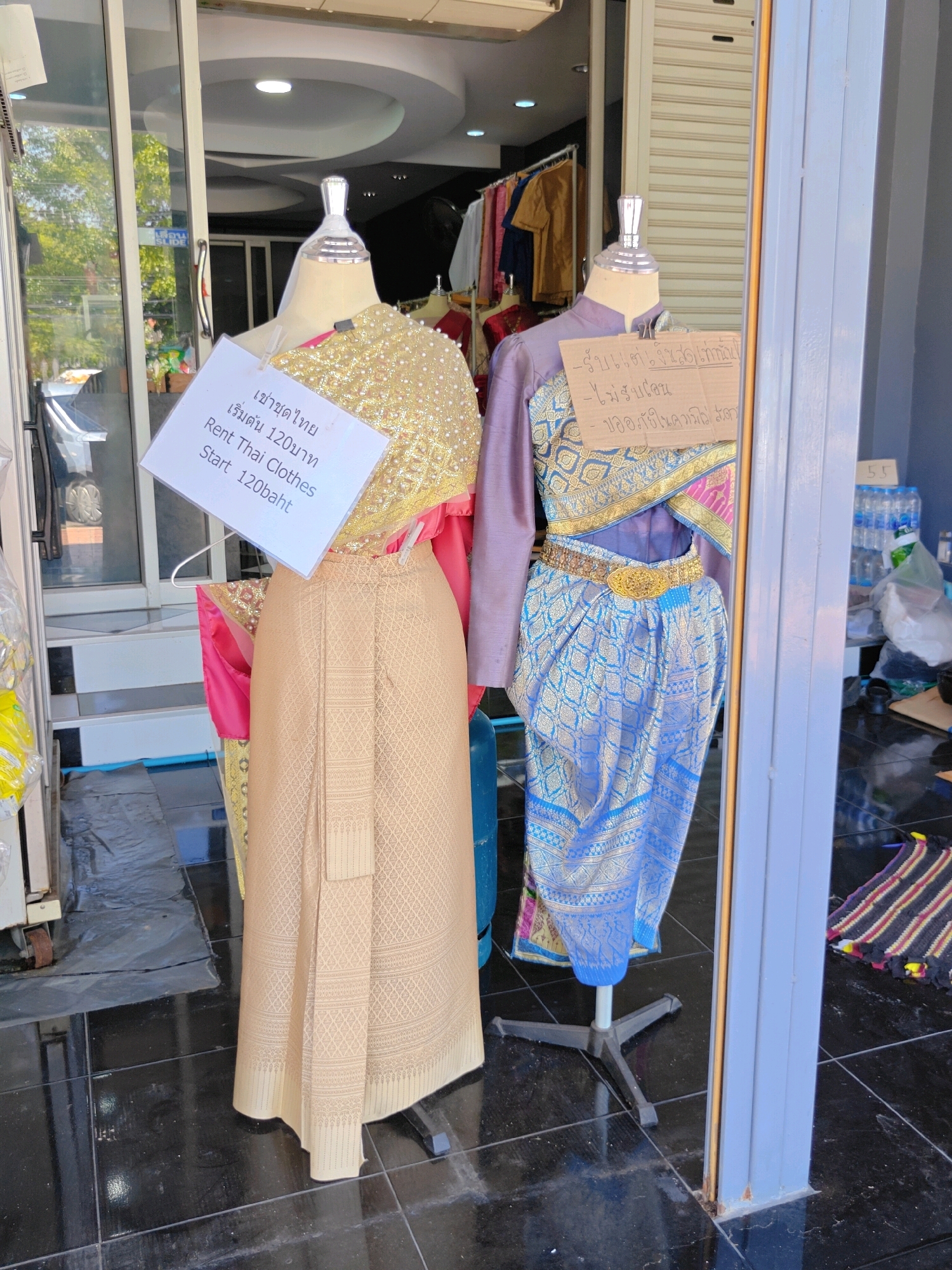 This is a medium shot, eye-level, outdoor photograph taken in Phra Nakhon Si Ayutthaya City Municipality, Thailand. The image features two mannequins dressed in traditional Thai clothing displayed in what appears to be the entrance of a shop.
The mannequin on the left is wearing a gold, heavily embellished top and a beige patterned skirt. A white sign is pinned to its chest, which reads in Thai and English: "เช่าชุดไทย / Rent Thai Clothes / เริ่มต้น 120บาท / Start 120baht".
The mannequin on the right is dressed in a purple top and a blue patterned skirt, adorned with a gold belt. A piece of cardboard is attached to its chest, with handwritten text in Thai which translates to: "-รับแต่ชุดไป... / -ไม่รับคืน / -ขอเก็บเงินให้ค่ะ / I only accept clothes to rent out. / No returns. / Please pay me."
The mannequins are positioned on a black tiled floor. In the background, through a glass door or window, clothing racks with various colorful garments are visible. To the right of the mannequins, a stack of bottled water and other items are stored, along with a small, colorful rug on the floor. The overall lighting suggests it is daytime, with sunlight casting shadows.