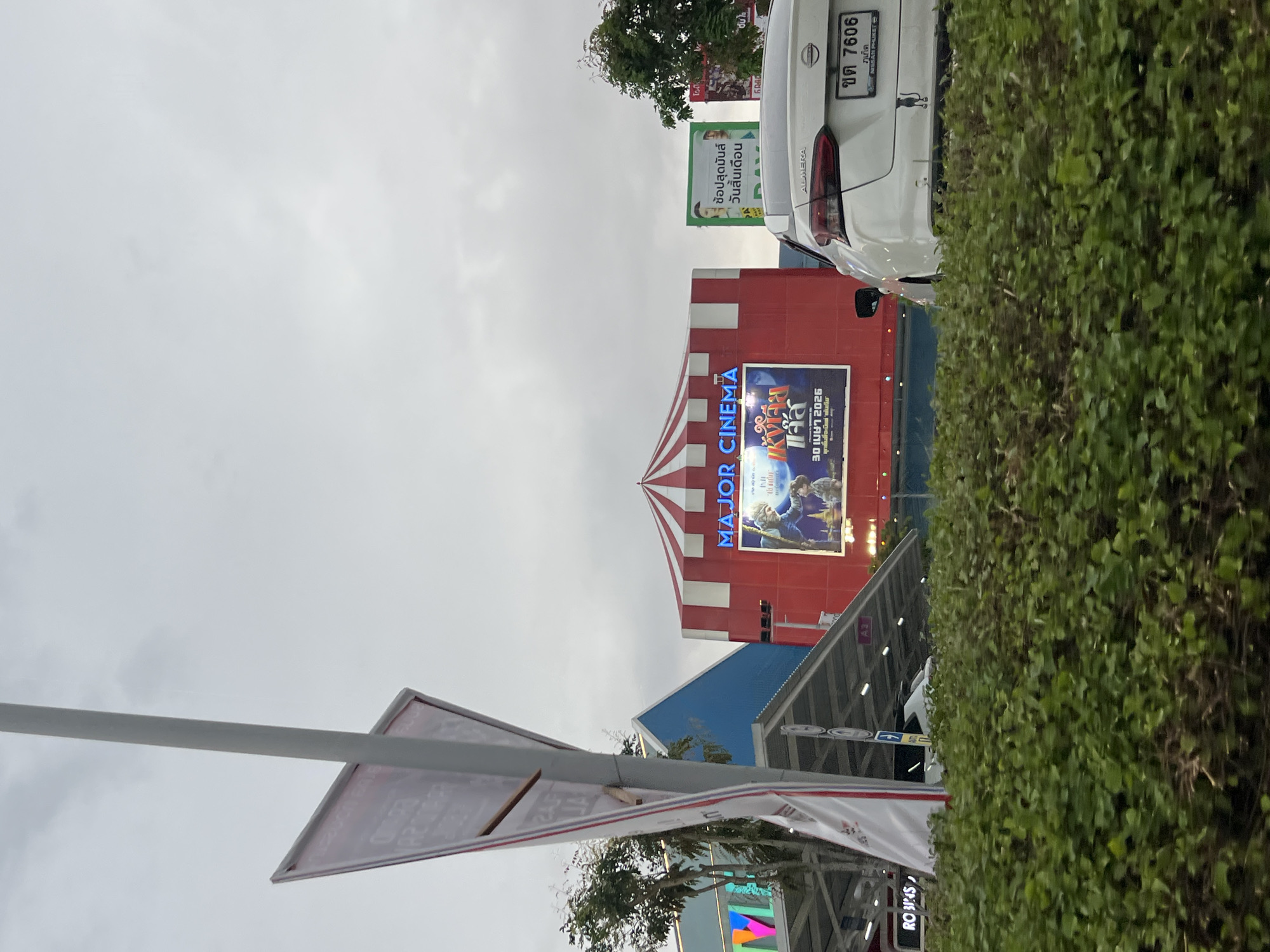 In Chalong, Thailand, a white Nissan Almera with license plate "ขต 7606" is parked in front of a Major Cinema. The cinema building features a distinctive red and white striped awning over its entrance. A large movie poster is displayed on the cinema's facade, advertising a film with Thai text. To the left of the cinema, a blue building and a section of a shopping complex are visible. In the foreground, a lush green hedge lines the roadway. The sky above is overcast.