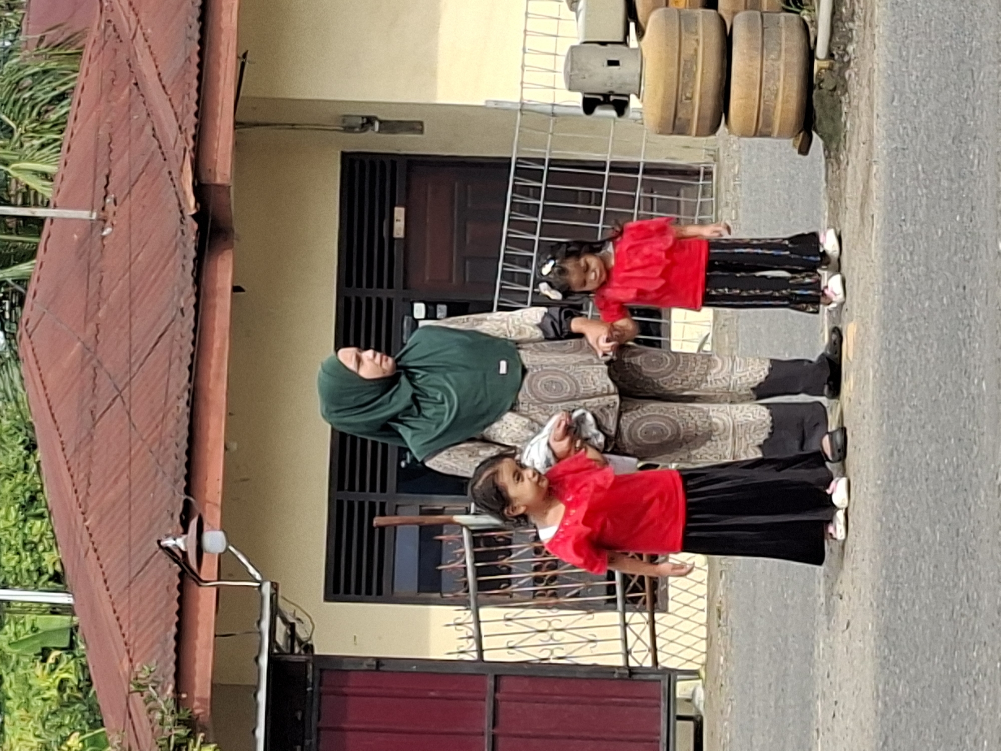 In Lhokseumawe, Indonesia, a woman wearing a green hijab and patterned clothing stands outdoors with two young girls. The girl on the left wears a red shirt and a long black skirt, while the girl on the right wears a red shirt and striped pants. The woman holds the hands of both children. They are standing on a paved road in front of a building with a red, corrugated metal roof and cream-colored walls. A metal fence is visible behind them, and to the right, there are stacked cylindrical objects. The environment appears to be residential or commercial, with trees visible in the background.