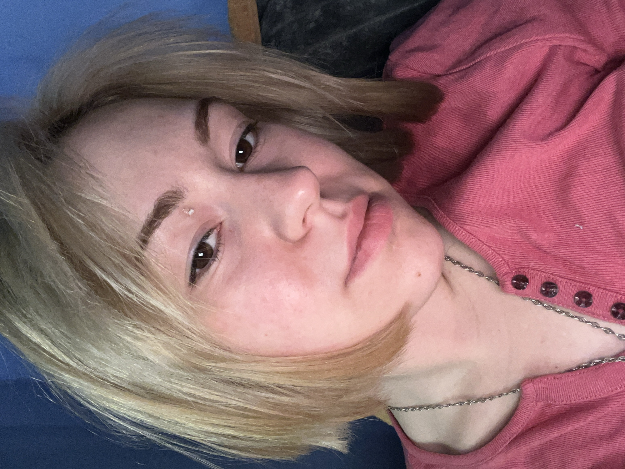 A close-up, angled portrait of a young individual, likely female, captured indoors in Odesa, Ukraine. The subject has fair skin, short-to-medium length blonde hair with visible darker roots, and dark brown eyes. She looks directly at the camera with a neutral to slightly serious expression. Notable facial features include natural skin texture, faint freckles or moles, and a small blemish or piercing on her forehead above her left eyebrow. Her lips are naturally colored and slightly parted.
She is wearing a long-sleeved, pink top or sweater featuring a ribbed texture. The garment has a placket with several dark, round buttons visible on the right side of the frame. A thin silver chain necklace is worn around her neck, resting on her top. The background behind her is composed of a solid blue surface on the left, possibly a wall or furniture, and a darker, textured fabric or material behind her head on the right. The lighting suggests an indoor setting, likely under artificial light. There is no discernible activity beyond posing for the photo, and no text is visible in the image.