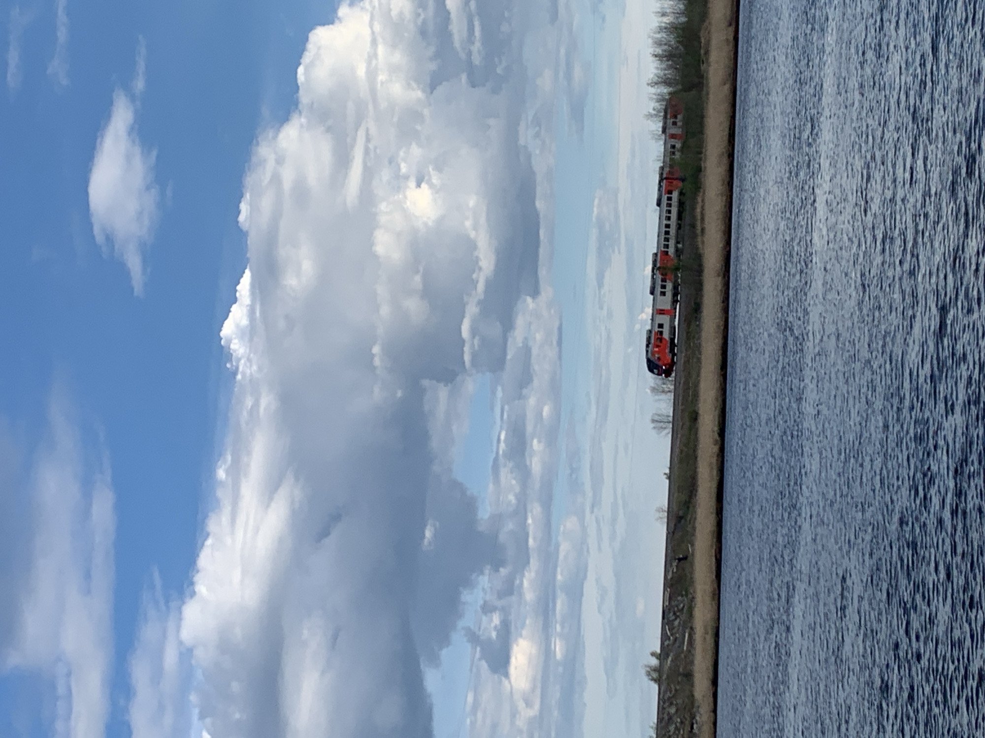 The image depicts a view of a body of water, possibly a lake or river, adjacent to a train track. A red and white train is visible on the track, which is bordered by a grassy area. The sky is dominated by white clouds. This scene could be in Severodvinsk, Russia, as it is a city known for its railway lines along bodies of water.