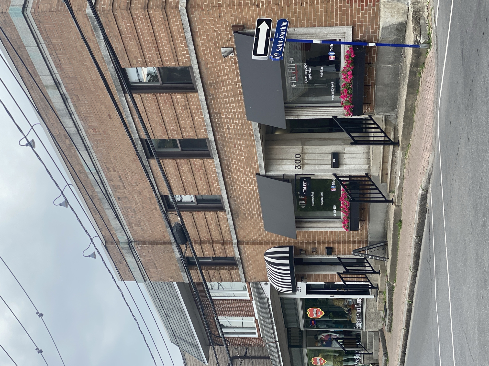This image shows the facade of a commercial building located on Saint-Joachim Avenue in Pointe-Claire, Canada. The building is constructed of red brick and features multiple storefronts. One storefront, labeled "TRI FIT," has a black awning and window signage indicating "Cours de groupe semi-privé" and "Nutrition," along with a website address and social media handles. Next to it, another storefront, labeled "296," has a black and white striped awning. Further to the left, a storefront displays mannequins in clothing, with signs reading "SpiceDaddy." A blue street sign indicates "Avenue Saint-Joachim" with a white directional arrow pointing right. Power lines are visible overhead. The street in front of the building is paved with asphalt and has white lane markings. A sidewalk, made of concrete and red pavers, runs along the building. Flower boxes filled with pink flowers are mounted on the facade of the "TRI FIT" establishment. The sky is overcast.