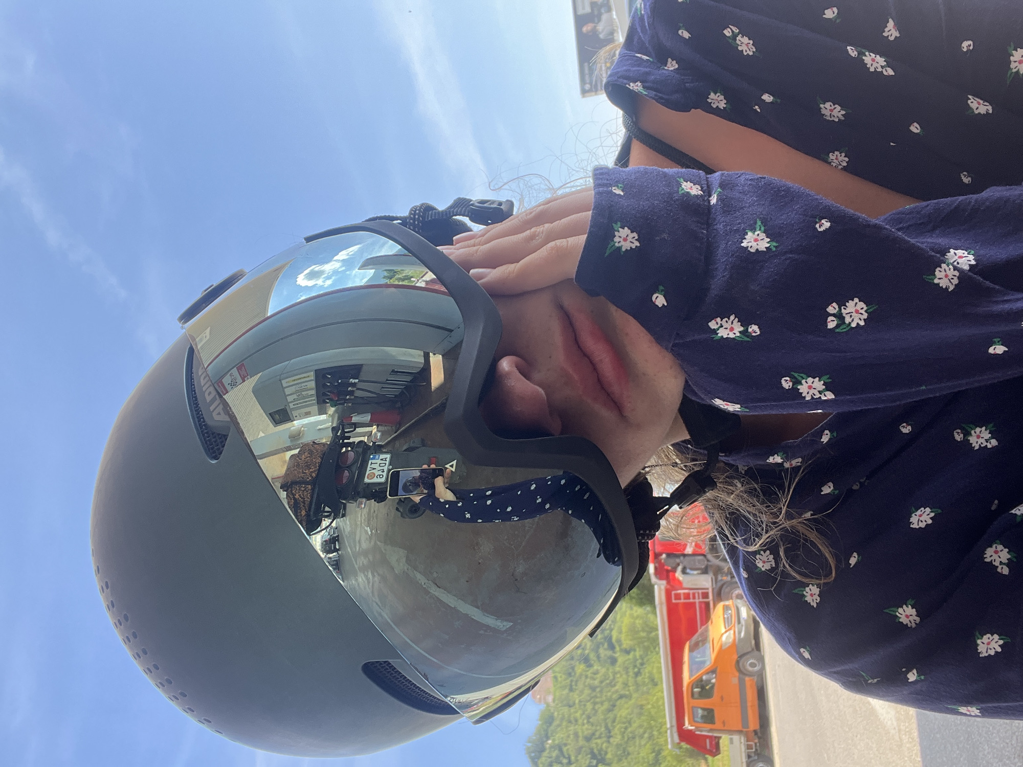 The image, rotated 90 degrees clockwise, portrays a person wearing a dark grey or black aerodynamic helmet with a highly reflective mirrored visor, standing outdoors under a bright blue sky. The person's hands are raised, covering their mouth and nose, which suggests shyness, playfulness, or surprise. They are dressed in a dark blue long-sleeved top adorned with small white and green floral patterns. Wisps of light-colored, curly hair are visible behind their ear and along their neck.
The polished visor of the helmet reflects the surrounding environment in detail. A clear reflection shows another person, possibly the same individual taking a selfie, holding a smartphone. Also visible in the reflection is a sign with the text "ALPINA" on a building, along with a red truck and an orange vehicle. A license plate, mirrored and upside down, displaying "VT 3ДОА" is prominently reflected on the visor. Green trees and parts of a bright sky are also seen in the reflection.
Beyond the helmet's reflection, the background features a clear blue sky with sparse white clouds on the left side of the frame. On the right, parts of a red truck and an orange vehicle are visible, corresponding to those seen in the visor's reflection. Green foliage from trees completes the outdoor setting. The scene is illuminated by bright daylight, indicating clear and sunny weather, likely taking place in Kubasi, Montenegro.