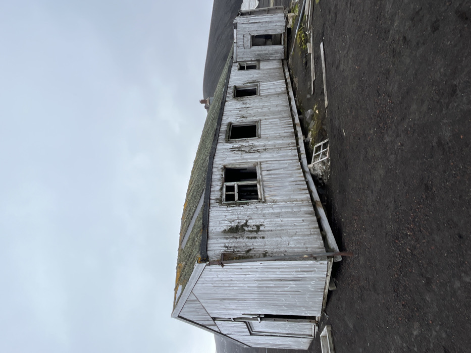A weathered, dilapidated white building stands against a gray sky. The building features a corrugated roof covered in moss, and peeling paint on its wooden siding. Several windows are open or missing panes, adding to the sense of decay. The ground is a dark, rough terrain, possibly dirt or gravel. The overall scene evokes a sense of abandonment and age.