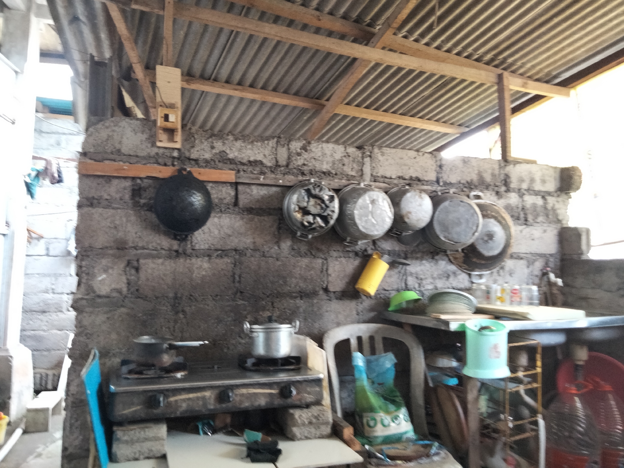 This is an interior shot of what appears to be an outdoor or semi-outdoor kitchen area in Denpasar, Indonesia. The walls are constructed from rough, gray cinder blocks, and the ceiling is made of corrugated metal supported by wooden beams. Hanging on the wall are various cooking pots, including a large black pot to the left and several smaller metal pots and pans.
In the foreground, a two-burner gas stove is visible, with a small pot on the left burner. To the right of the stove, a white plastic chair is positioned, and a blue and green bag with a white pattern rests on it. A metal shelf unit is situated further to the right, holding several items including a mint green container, stacked plates, and clear glass bottles. A large red basin or bowl is also visible on the right side of the image. The overall impression is of a functional but rustic kitchen space.