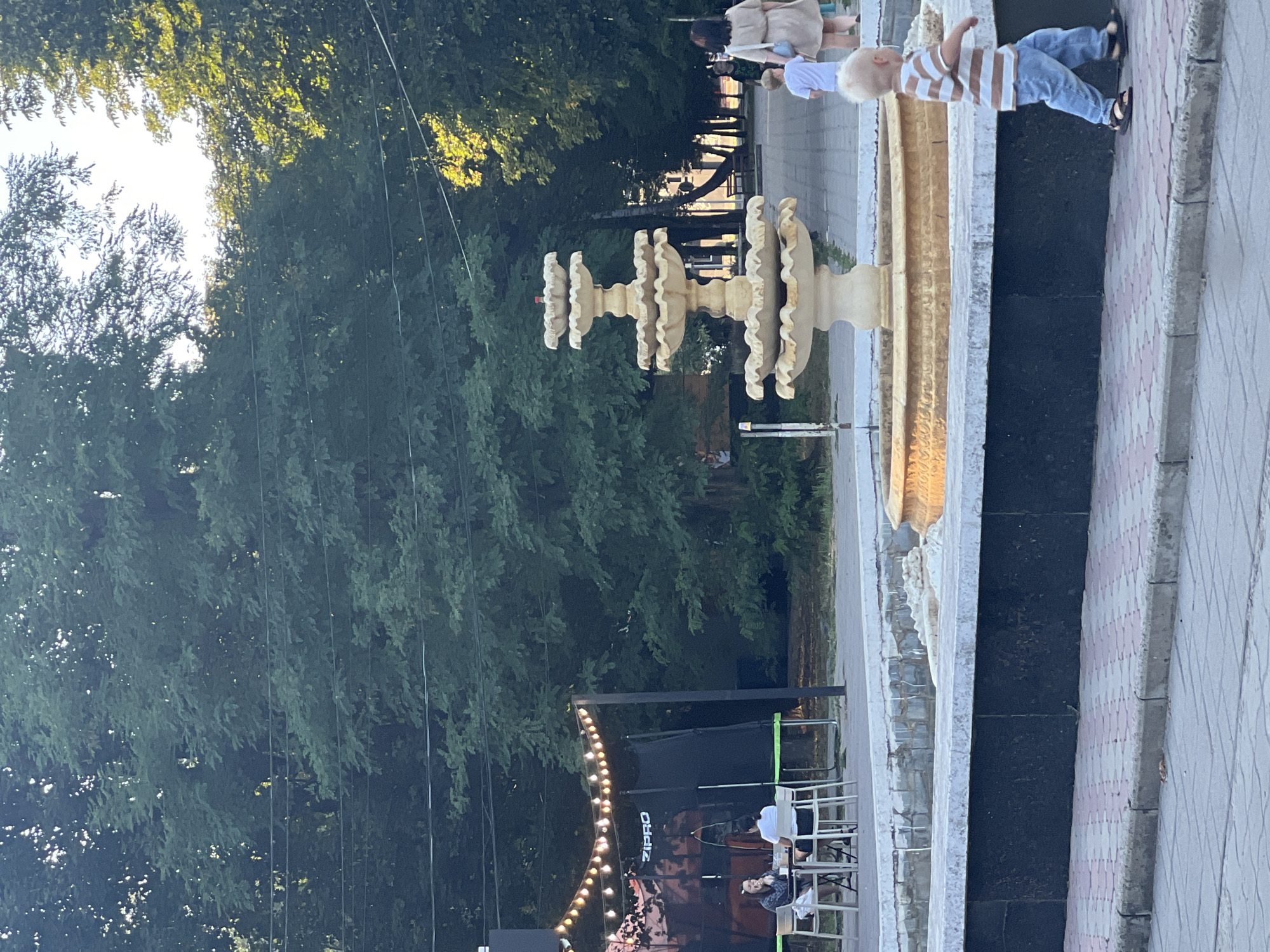 In the city of Sheptytskyi, Ukraine, a park scene unfolds with a tiered fountain as the central focus. The fountain, made of a light-colored material, is surrounded by a dark, tiled base and a white stone ledge. To the left, two women are seated at a table, with string lights illuminating the area behind them and a black trampoline visible. Further back, several people are walking along a path, including a woman in a light-colored dress and a young boy in a striped shirt and blue jeans. The environment is lush with mature trees providing shade, and power lines are visible strung between them. The ground is paved with a patterned brick walkway.