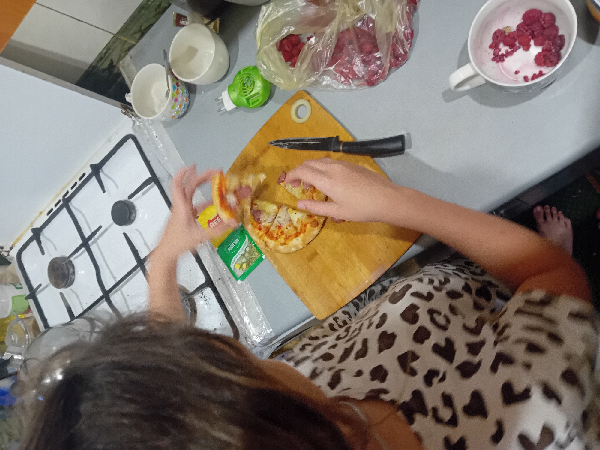 The image depicts a domestic scene, likely a kitchen, where a young person is preparing food. A small pizza, cut into slices, rests on a wooden cutting board. The person, seen from the shoulders down, is in the process of handling a slice of the pizza. A knife is also on the cutting board. Nearby, a plastic bag contains raspberries, and a white mug holds more raspberries. Other objects visible include a gas stove with burners, several white cups, and a small green object that appears to be an electrical plug. The overall setting suggests a casual, home-cooked meal preparation. There is no information in the image that indicates the location is in the city of Chuhuiv, Ukraine.