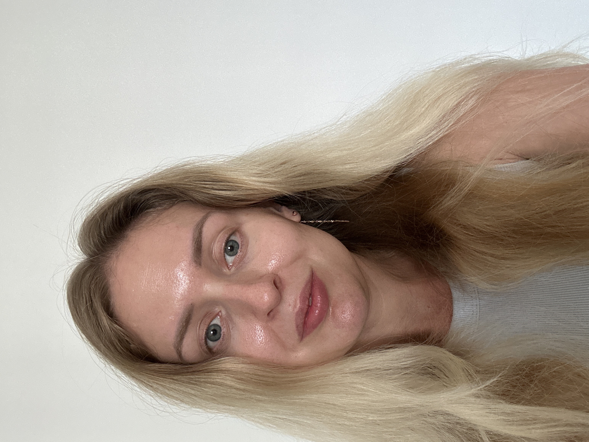 A fair-skinned individual with long, blonde hair and blue eyes is depicted in a headshot, looking directly at the camera with a slight smile. The subject's forehead, nose, and cheeks exhibit a prominent sheen, suggesting a dewy or oily complexion. A delicate, dangling earring is visible in the subject's right ear. The individual is wearing a light-colored, ribbed top. The background is an undifferentiated, plain white wall. The image contains no visual cues or specific details to confirm the stated location of Petah Tikva, Israel.