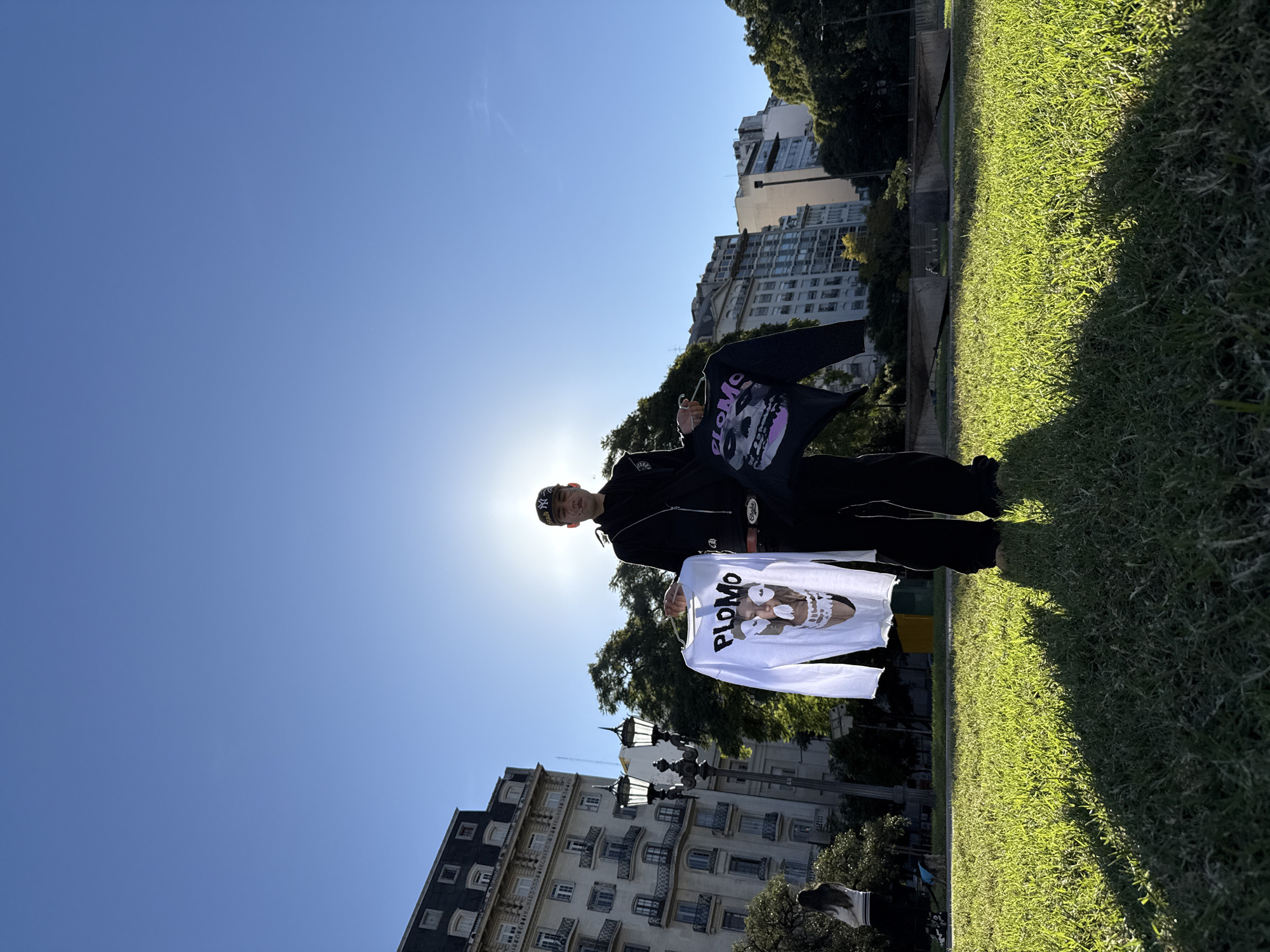 A full-length shot, from a low angle, portrays a person standing on a grassy lawn in what appears to be a park or green area in Buenos Aires, Argentina. The subject is holding two pieces of clothing: a white long-sleeved shirt with the word "PLOMO" and a graphic image on the front, and a dark-colored garment with a similar design. The person is wearing a dark tracksuit, cap, and sneakers. Buildings are visible in the background, indicating an urban environment, with one appearing to be a stately apartment building with multiple balconies. Street lamps are also visible. The image is taken during the daytime, with a bright sun creating a lens flare effect. The photo is rotated, displaying the sky on the left and grass on the right.