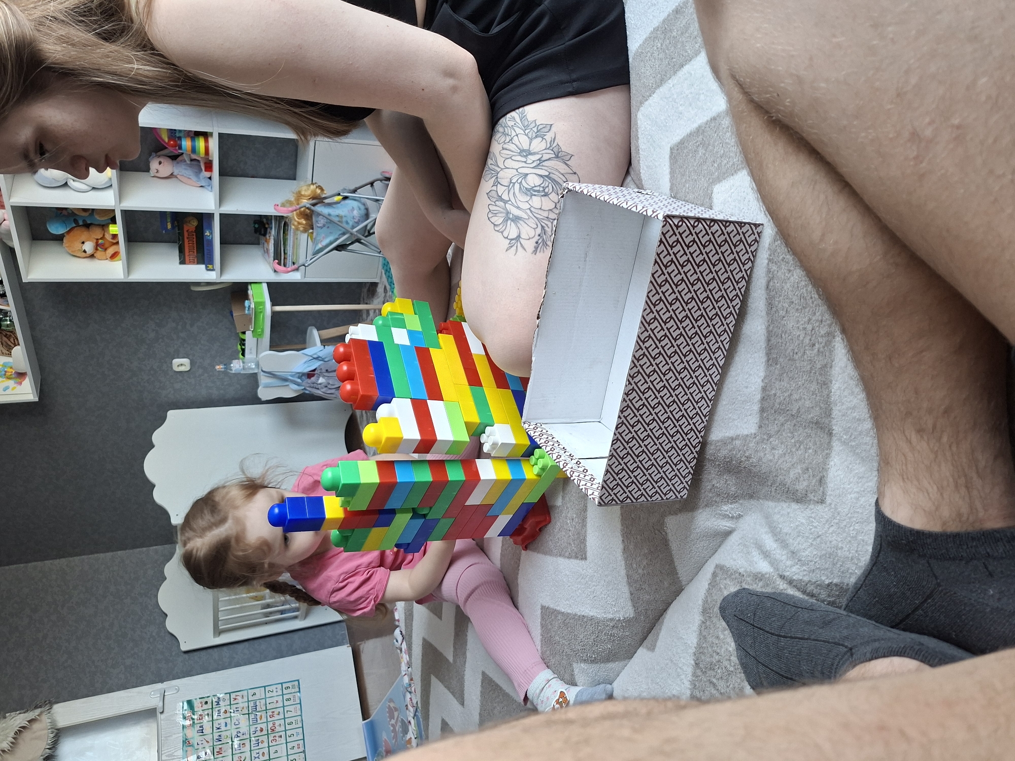 The image shows a scene within a home. A child wearing a pink shirt and light pink leggings is holding a large, multi-colored toy made of building blocks. A woman with light brown hair, wearing black shorts and with a tattoo on her thigh, is seated nearby, partially visible in the upper portion of the frame. Next to her is a patterned box. The scene is set on a patterned rug or blanket on the floor. In the background, a white shelf unit with various toys and objects is visible, along with a white changing table. A person wearing grey socks and having hairy legs is also in the shot.