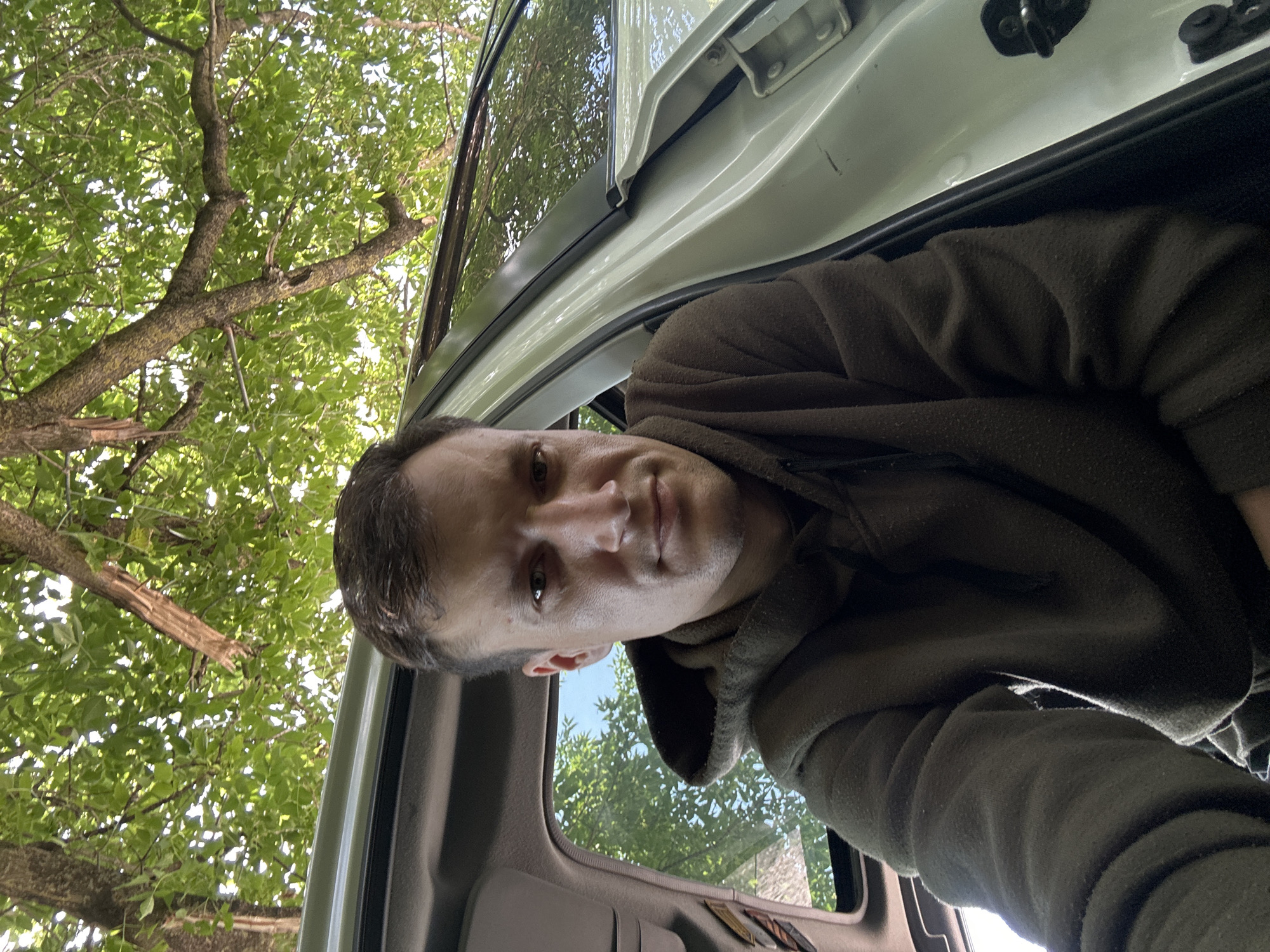 A young man with short, dark hair and fair skin is seated inside a vehicle. He is wearing a dark olive green hooded sweatshirt. The man is looking directly at the camera with a neutral expression. The vehicle's interior is visible, including a light-colored door panel and window frame. Outside the vehicle, green foliage from trees hangs over the scene, suggesting an outdoor setting, possibly a park or roadside area. The lighting indicates it is daytime. The exact location in the city of Makiv, Ukraine, cannot be determined from the image alone.