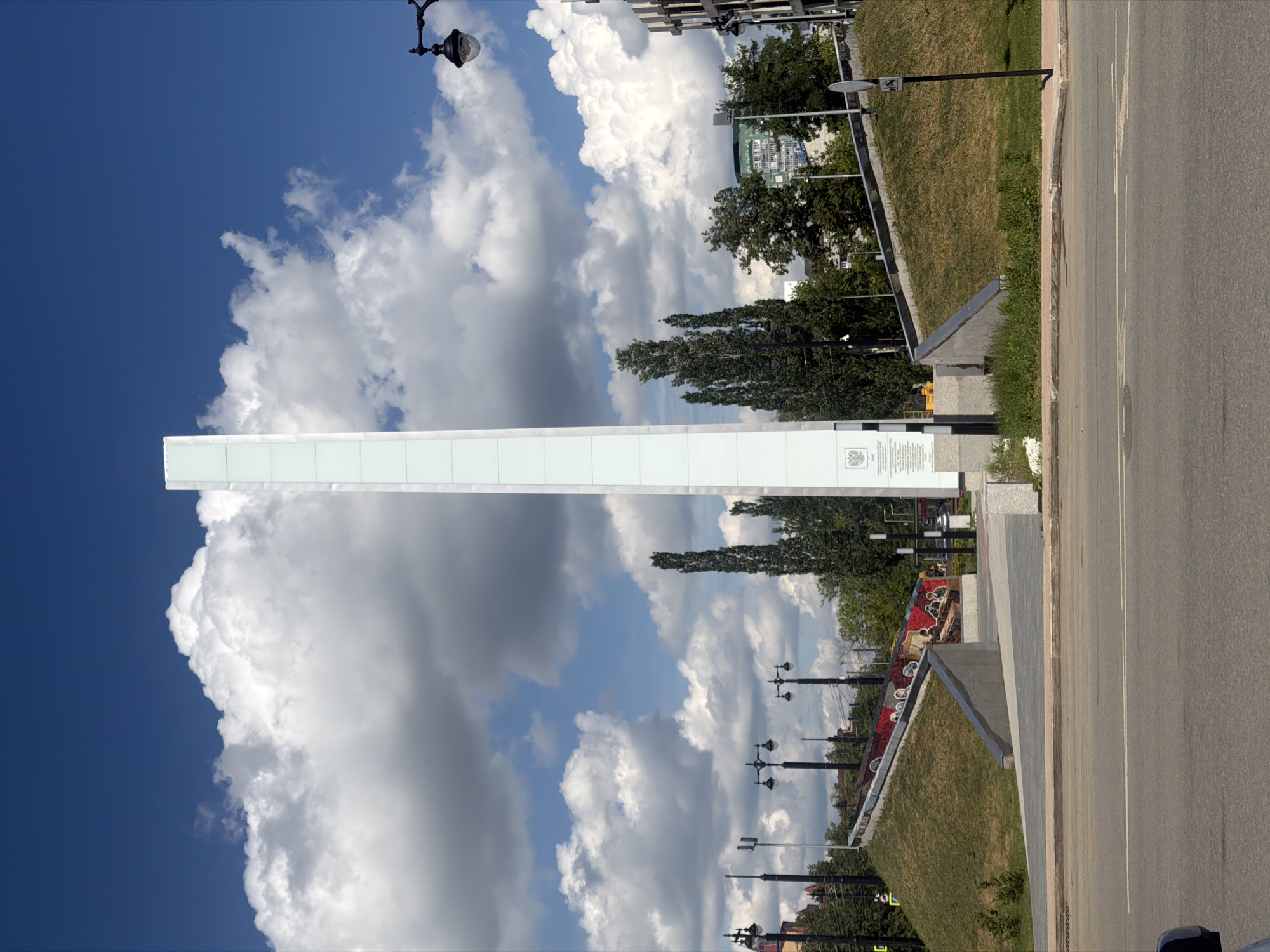 The image shows the "Wings of Victory" monument in Perm, Russia. The monument is a tall, white, rectangular structure, reaching towards the sky, which is filled with cumulus clouds against a blue background. To the right of the monument, there is a view of a road, a grassy slope, trees, street lamps, and some buildings in the distance. Some construction seems to be in progress on the grass slope as there is red and white marking tape and construction equipment.