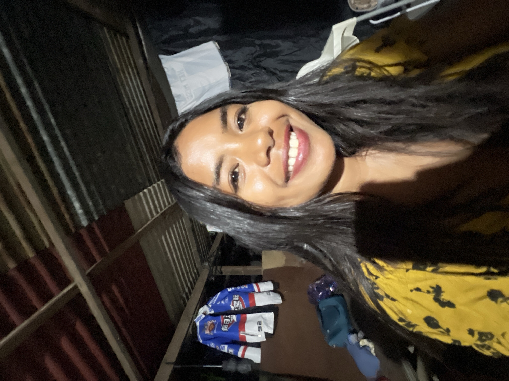 The image captures a young woman with long, dark hair and a bright smile, taking a selfie in an outdoor, likely informal, setting. She is wearing a yellow top with a black floral pattern. Behind her, a blue and white racing jacket with Japanese characters is hanging on a wooden beam. The environment appears to be a covered area with a corrugated metal roof and exposed wooden beams, possibly a patio, workshop, or an unfinished structure. The background is dark, suggesting it is either evening or nighttime. The overall impression is a casual and candid moment.