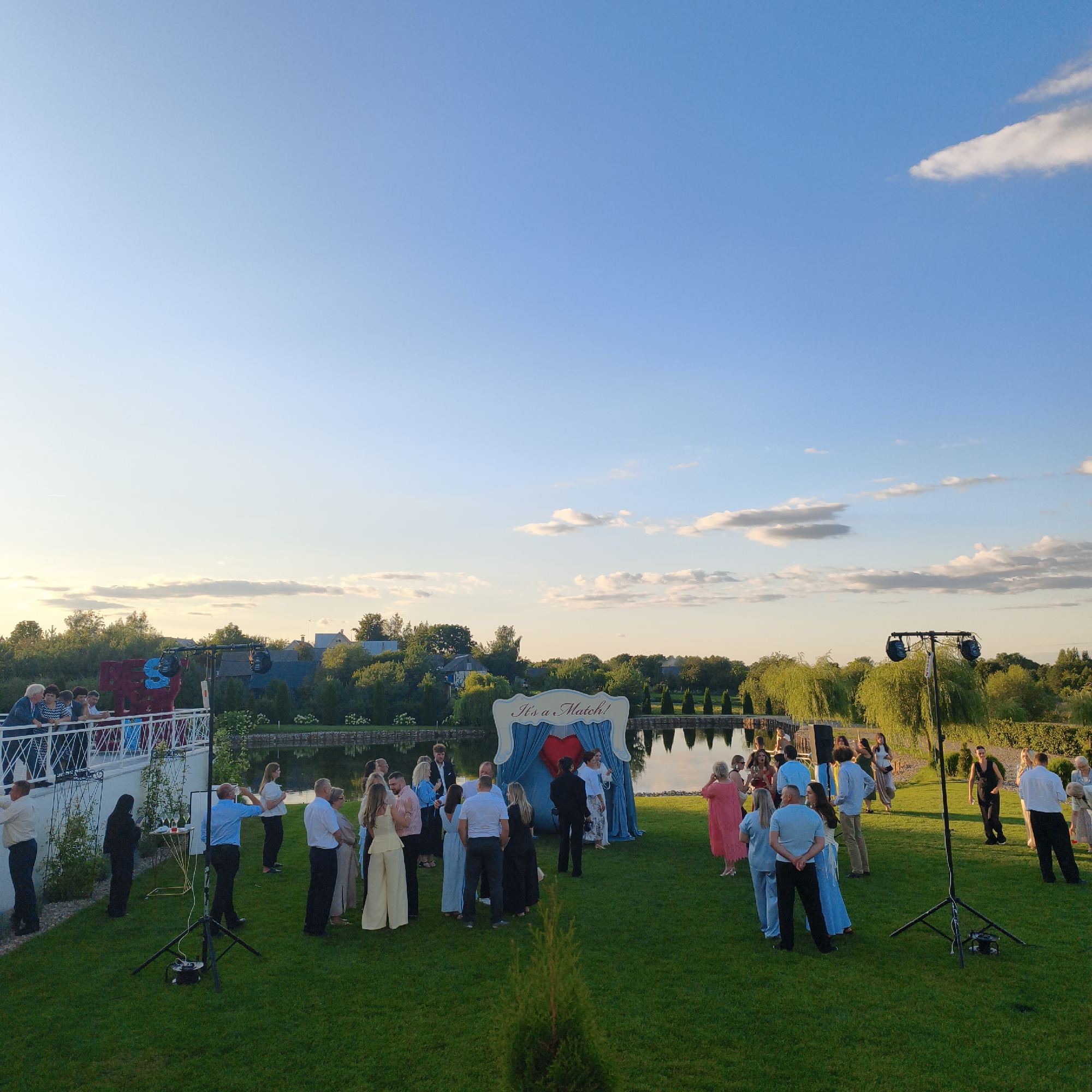 This image depicts an outdoor gathering in the Сапоцкінскі сельскі Савет, Belarus, likely a wedding reception or similar event, set against a backdrop of a serene lake and lush greenery. The sky is a clear, bright blue with a few wispy clouds, and the late afternoon sun casts a warm glow on the scene.
In the foreground, a group of people are mingling on a meticulously manicured lawn. They are dressed in a variety of attire, ranging from casual to semi-formal. Several individuals are standing and conversing, while others are seated or engaged in activities. A prominent element in the middle ground is a decorated archway with the words "It's a Match!" written above it, adorned with blue drapes and a red heart. This archway is situated near the edge of the lake, where trees with drooping branches line the shore, their reflections visible in the calm water.
To the left of the scene, a raised white platform or balcony overlooks the gathering, with several people standing and observing. On the right side, a tall light stand with two spotlights is positioned, suggesting that the event may extend into the evening. Scattered across the lawn are various other objects, including a small table with drinks, portable lighting equipment, and a young evergreen tree. The overall impression is one of a joyous and celebratory occasion in a picturesque natural setting.