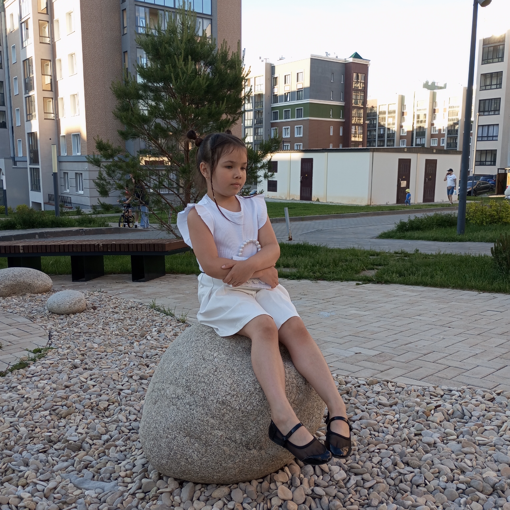 In the city of Pervomaysky, Russia, a young girl with dark hair styled in two buns is seated on a large, rounded grey boulder. She is wearing a white sleeveless top with ruffled shoulders and white shorts or a skirt. Her arms are crossed in front of her, and she looks to her left with a neutral expression. She is wearing black mesh flats with ankle straps. The boulder is surrounded by a bed of small, light-colored pebbles. Behind the girl, a wooden bench with dark metal legs is visible, as well as a paved walkway with a herringbone pattern. In the background, several modern apartment buildings with varying architectural styles and colors are present, some with balconies and large windows. A tall evergreen tree stands between the girl and the buildings. Further in the background, a man in a white t-shirt and blue shorts is talking on a phone, and another small child in a blue outfit and yellow hat is walking. Several cars are parked near the buildings. The scene is illuminated by natural sunlight.