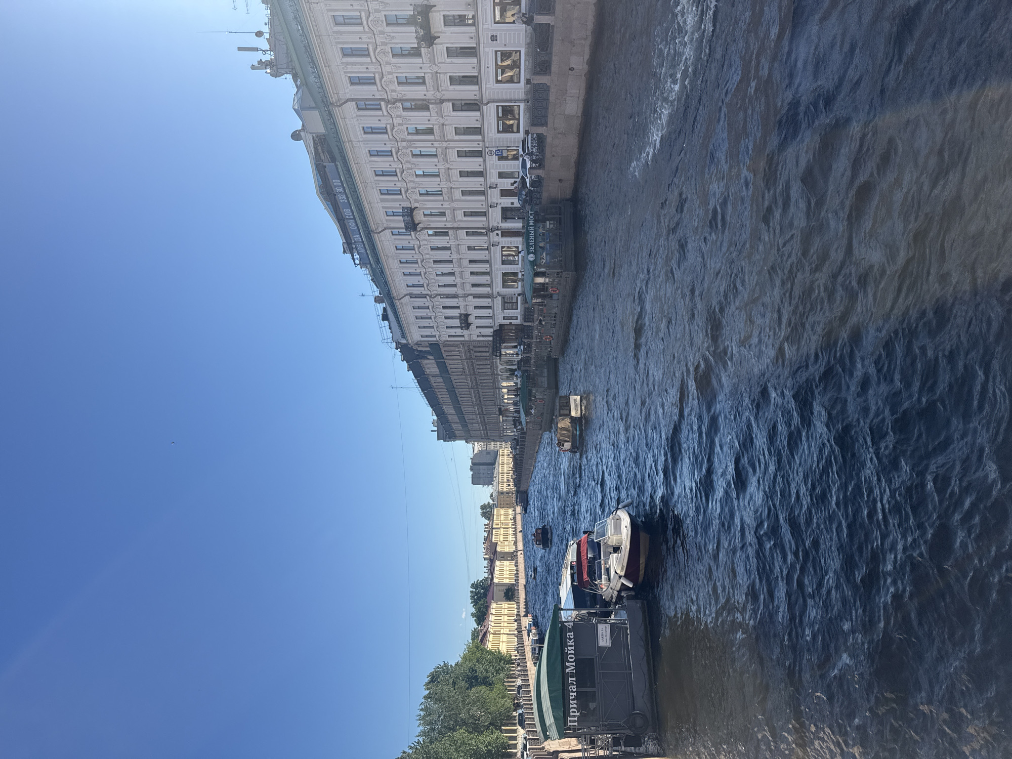 The image depicts a sunny day in Saint Petersburg, Russia, likely taken from a bridge. A wide canal, possibly the Moyka River, flows through the scene, with several tour boats and smaller vessels navigating its dark blue, rippling water. The canal banks are lined with ornate, historic buildings, characterized by their pastel facades, numerous windows, and detailed architectural elements. Trees with lush green foliage are visible on the left bank, adding to the picturesque urban landscape. On the right bank, a prominent building exhibits a row of windows and balconies, with some signage visible, including what appears to be an advertisement for office rentals. The sky is a clear, bright blue, with the sun casting a gentle light on the scene. The overall impression is one of a vibrant, historical city on a beautiful summer day.