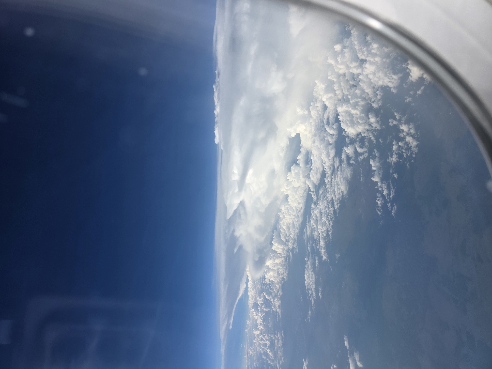 An aerial view from an airplane window shows a vast expanse of clouds under a deep blue sky. The clouds are layered, with some appearing as fluffy cumulus formations closer to the ground, and a large, flat expanse of stratus clouds stretching across the horizon. The sunlight illuminates the tops of the clouds, creating bright white highlights against the darker undersides and the deep blue of the sky. The window frame is visible in the foreground, a silver-toned metallic structure. The scene appears to be over a rural or less populated area, as no urban structures or clear land features are discernible. The image does not depict any people, objects, or actions, and its location in Khimki, Russia, cannot be confirmed from the visual information alone.