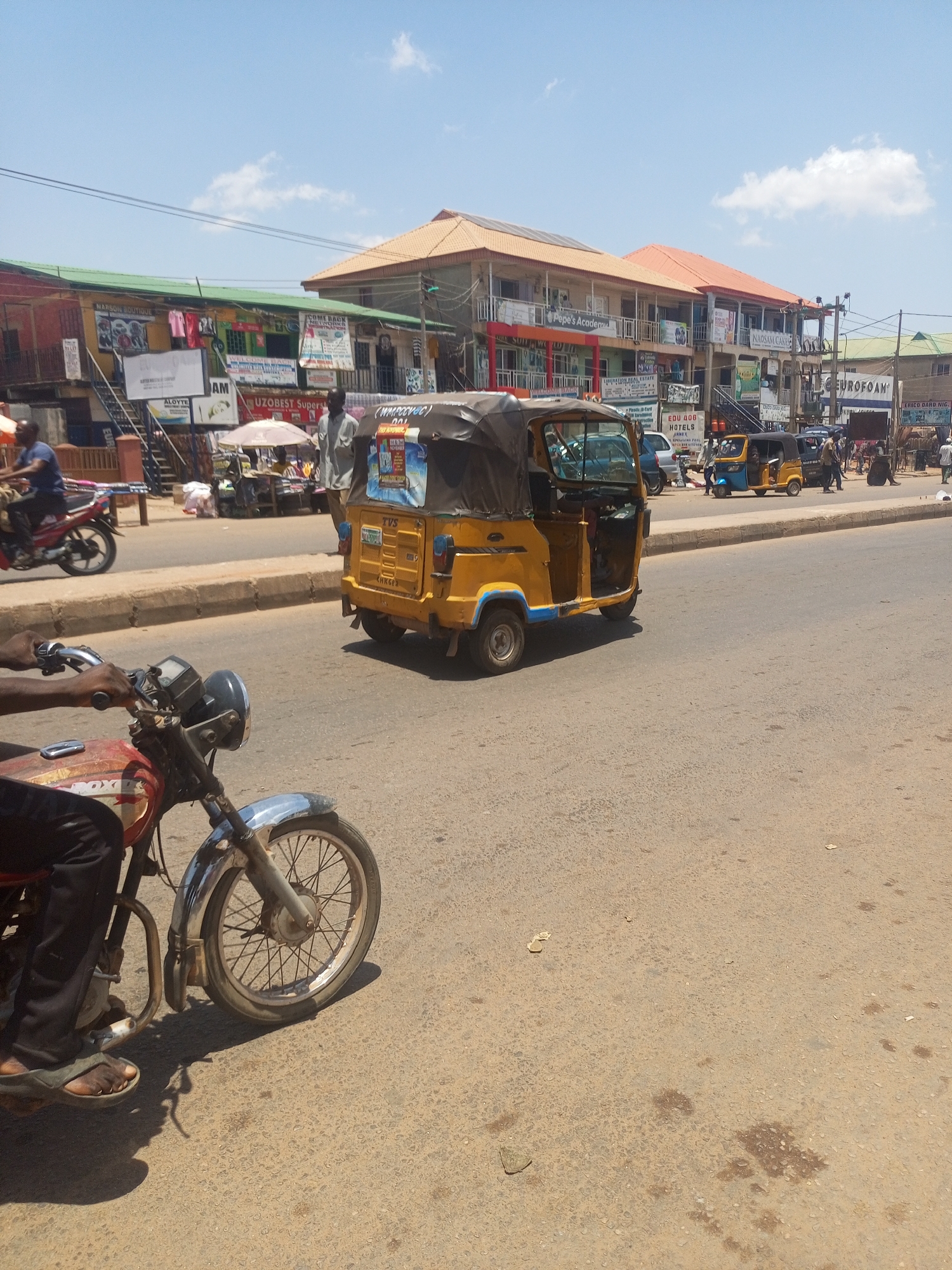The image depicts a street scene in Sabon Tasha, Nigeria, characterized by urban activity and a mix of commercial establishments. The street is paved and shows signs of wear. A yellow tricycle taxi is the central subject, with a motorcycle driven by a person wearing sandals in the foreground. Another motorcycle is visible in the background. Multi-story buildings line the street, displaying various business names, including "NARSON BOUTIQUE," "UZOBEST Supers," "Pepe's Academic," "EDU 406 HOTELS," "NAOSAM CAKES," and "EUROFOAM." Vendors and pedestrians are present along the sidewalk. The sky is blue with scattered clouds. The overall scene conveys a typical busy day in an urban setting in Nigeria.