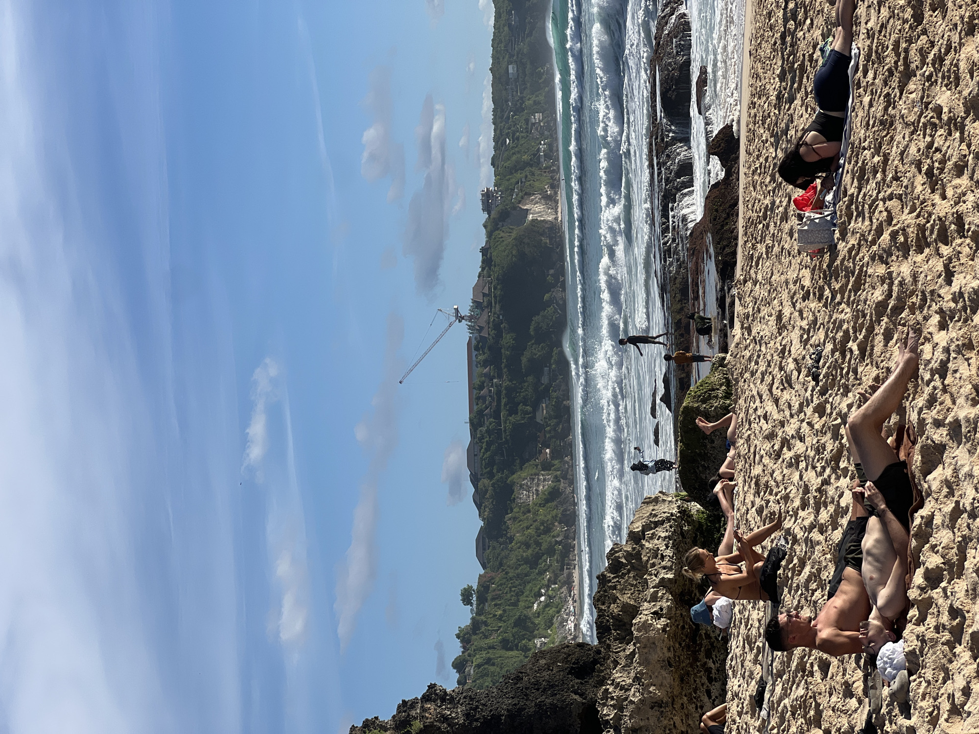The image depicts a beach scene in Kuta Selatan, Indonesia. People are relaxing on the sand, some sunbathing, near the ocean. The coastline is visible with waves crashing. A crane is present on the shoreline, indicating construction or development in the area. The sky is blue with some clouds.