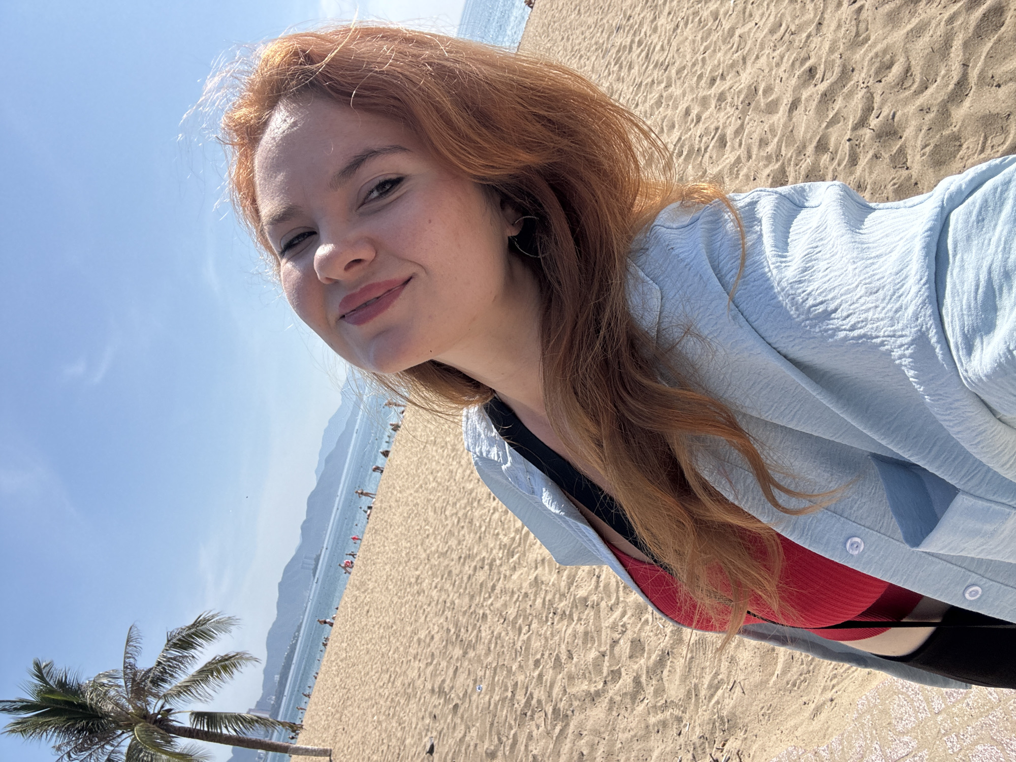 In Nha Trang Ward, Vietnam, a young woman with red hair takes a selfie on a sandy beach under a clear blue sky. She is smiling and looking towards the camera. She is wearing a red top and a light blue, textured shirt open at the front. In the background, the ocean stretches out, dotted with distant mountains and a palm tree to the left. Other beachgoers are visible in the distance, enjoying the sunny day. The sand is golden and textured, with some subtle ripples.