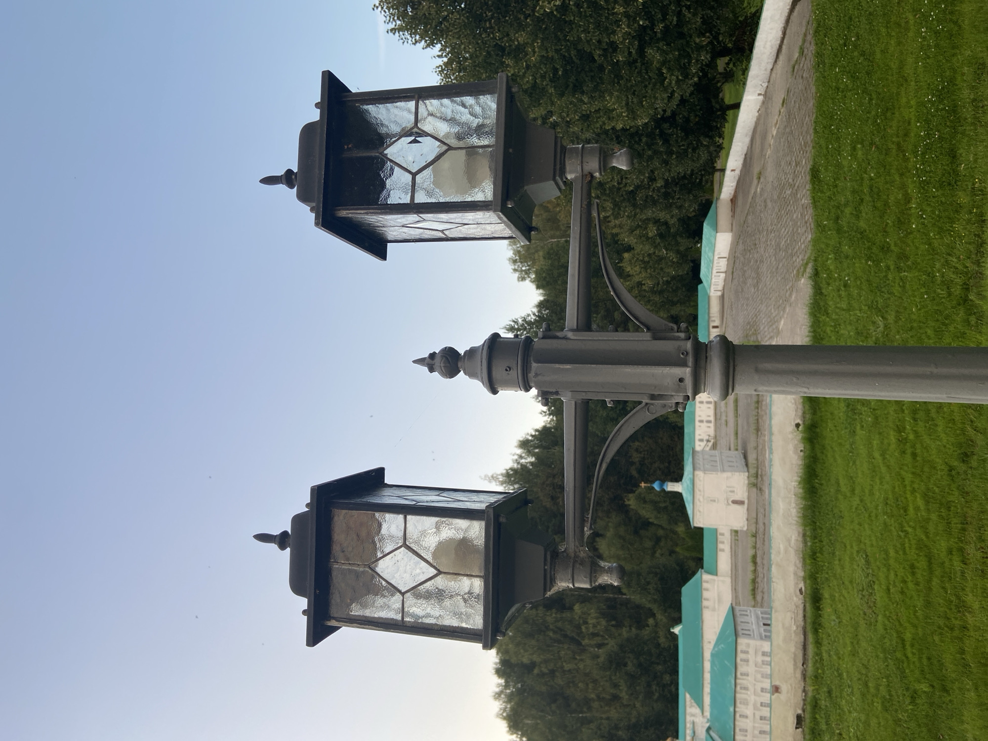 This is a photograph of a double lamp post with a weathered, dark grey metal finish, set against a pale blue sky. The lamp post has two ornate lamps with decorative finials. The lamps are hexagonal in shape with textured glass panels, each containing a light bulb. In the background, there are trees and a miniature village, possibly a model of a monastery. The village features white buildings with turquoise roofs, arranged along a cobblestone path. The foreground is a grassy lawn dotted with small white flowers. The scene appears to be outdoors in a park-like setting. The location is identified as the city of Chulis, Russia.