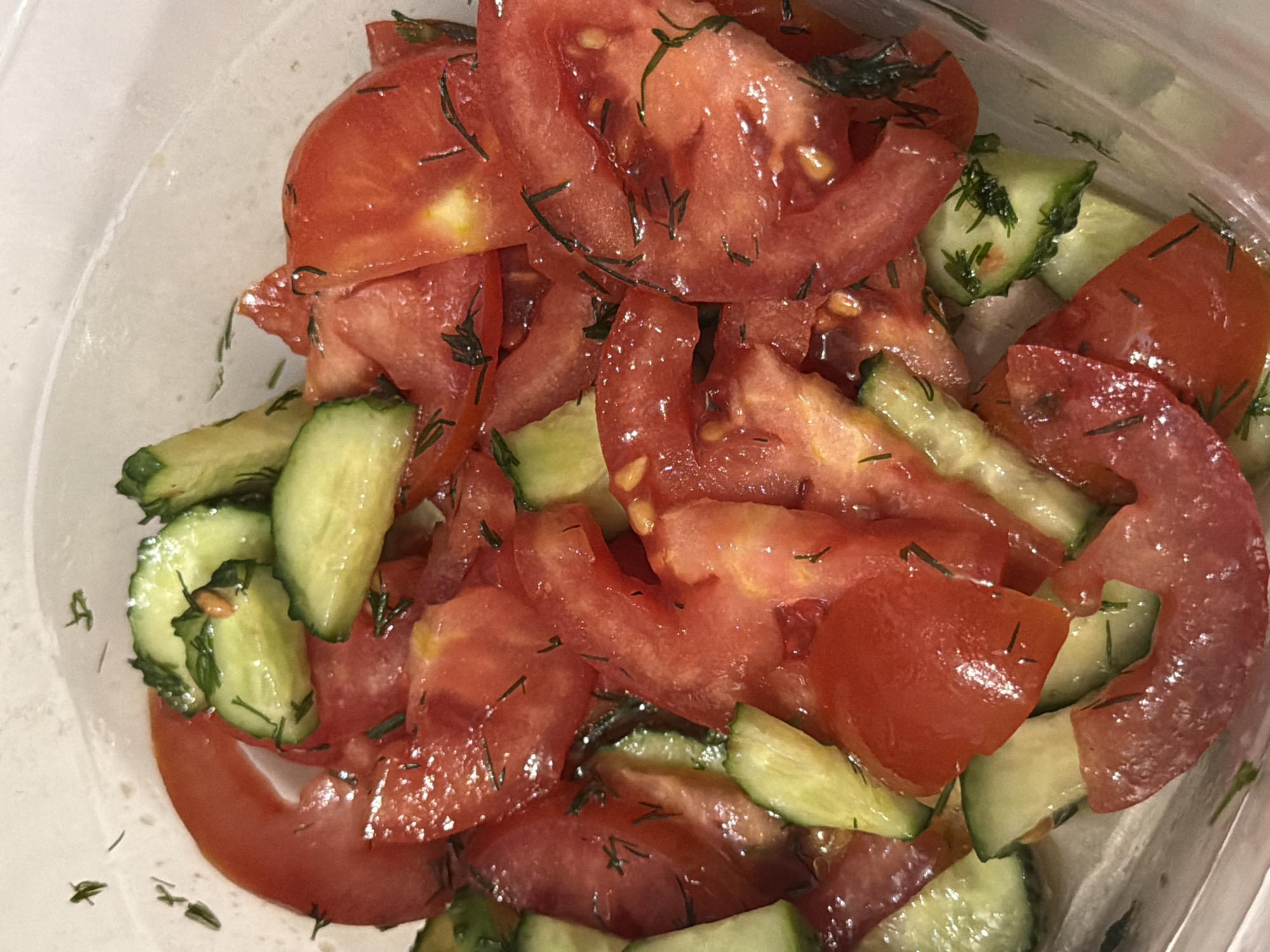 A close-up shot shows a clear plastic container filled with chopped tomatoes and cucumbers, seasoned with dill. The tomatoes are bright red and juicy, sliced into irregular pieces. The cucumbers are green and also chopped into various shapes. Tiny green dill sprigs are scattered throughout the salad, adding a fresh, herbaceous aroma. The lighting is bright, casting slight reflections on the moist ingredients. The background is out of focus, showing the translucent texture of the container. The scene appears to be an indoor setting, likely a kitchen or dining area.