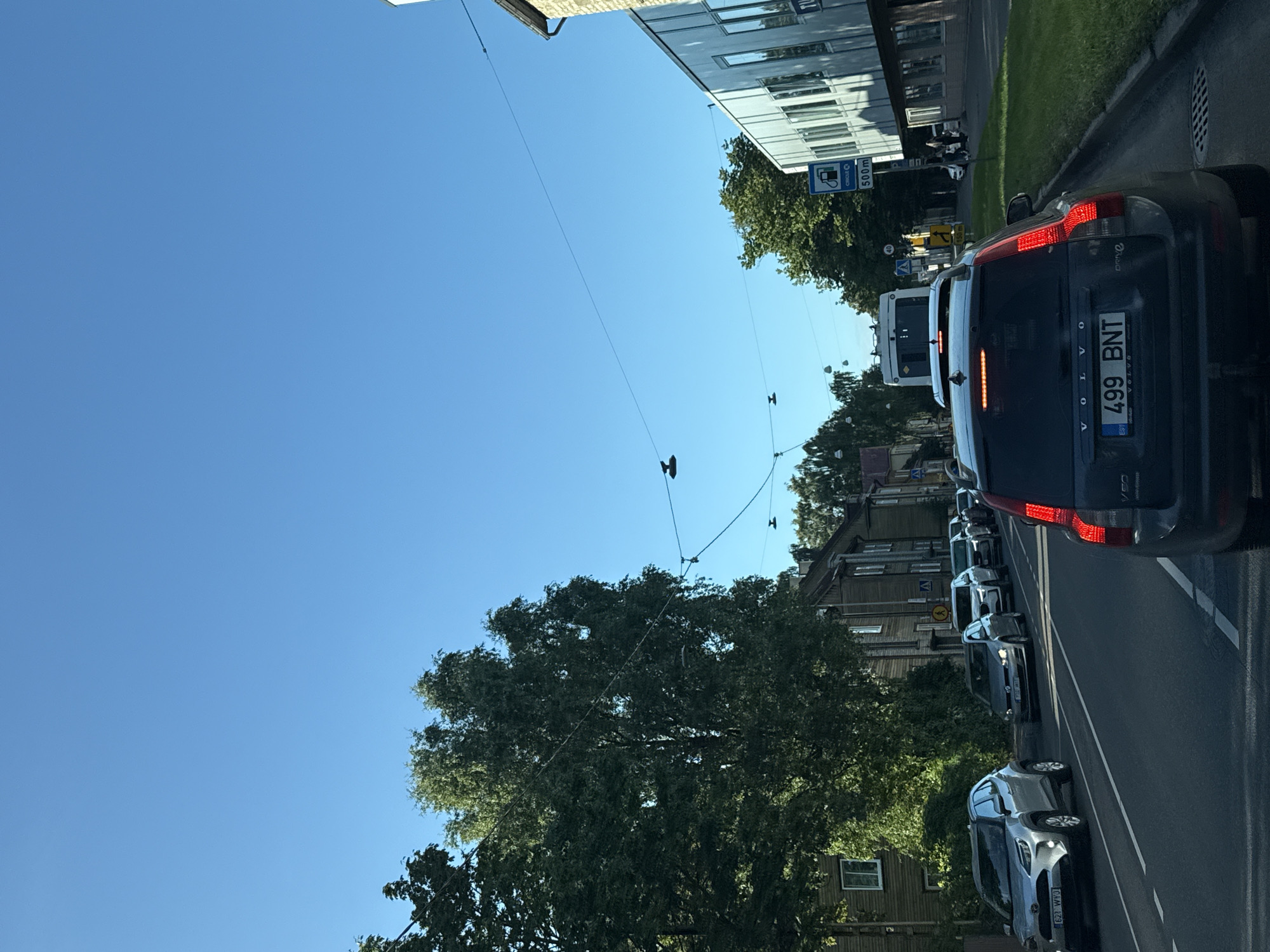 The image depicts a street scene in Tallinn, Estonia, on a clear, sunny day. The perspective is from inside a vehicle, looking forward. The road is lined with trees and buildings on either side.
On the left side of the road, several cars are visible, including a silver Toyota Yaris with license plate "621 WYJ". Further down the road, more vehicles are in traffic. The buildings on the left appear to be older, wooden structures.
On the right side of the road, a modern building with large windows is present. A blue sign indicating a "Circle K" gas station with "500m" distance is displayed. Traffic lights and street lamps are also visible.
In the foreground, a dark-colored Volvo station wagon is seen, with its rear lights illuminated and license plate "499 BNT". Several other vehicles are in front of it, forming a traffic queue. A bus is also present further up the road. The sky is a clear, bright blue. The overall scene suggests a typical urban environment with moderate traffic.