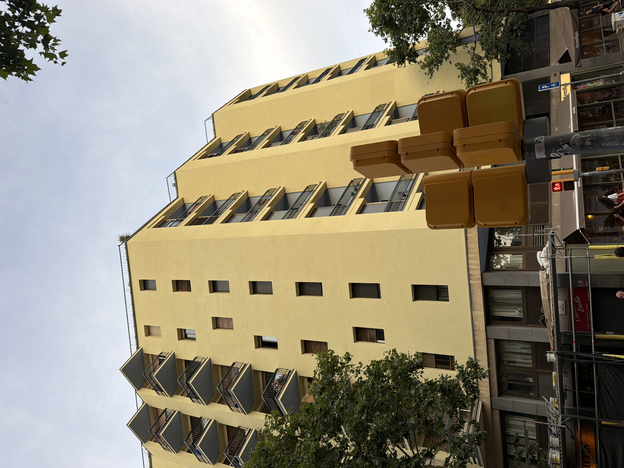 An exterior, low-angle, medium shot captures a tall, yellow, modern apartment building in Barcelona, Spain, under a cloudy sky. The building features multiple floors with balconies and windows. In the foreground, a traffic signal with yellow lights is prominently displayed. Trees with dark green foliage frame the building on the left and right sides. The bottom of the image shows a street-level view with scaffolding and awnings of storefronts. The scene appears to be captured during the daytime.