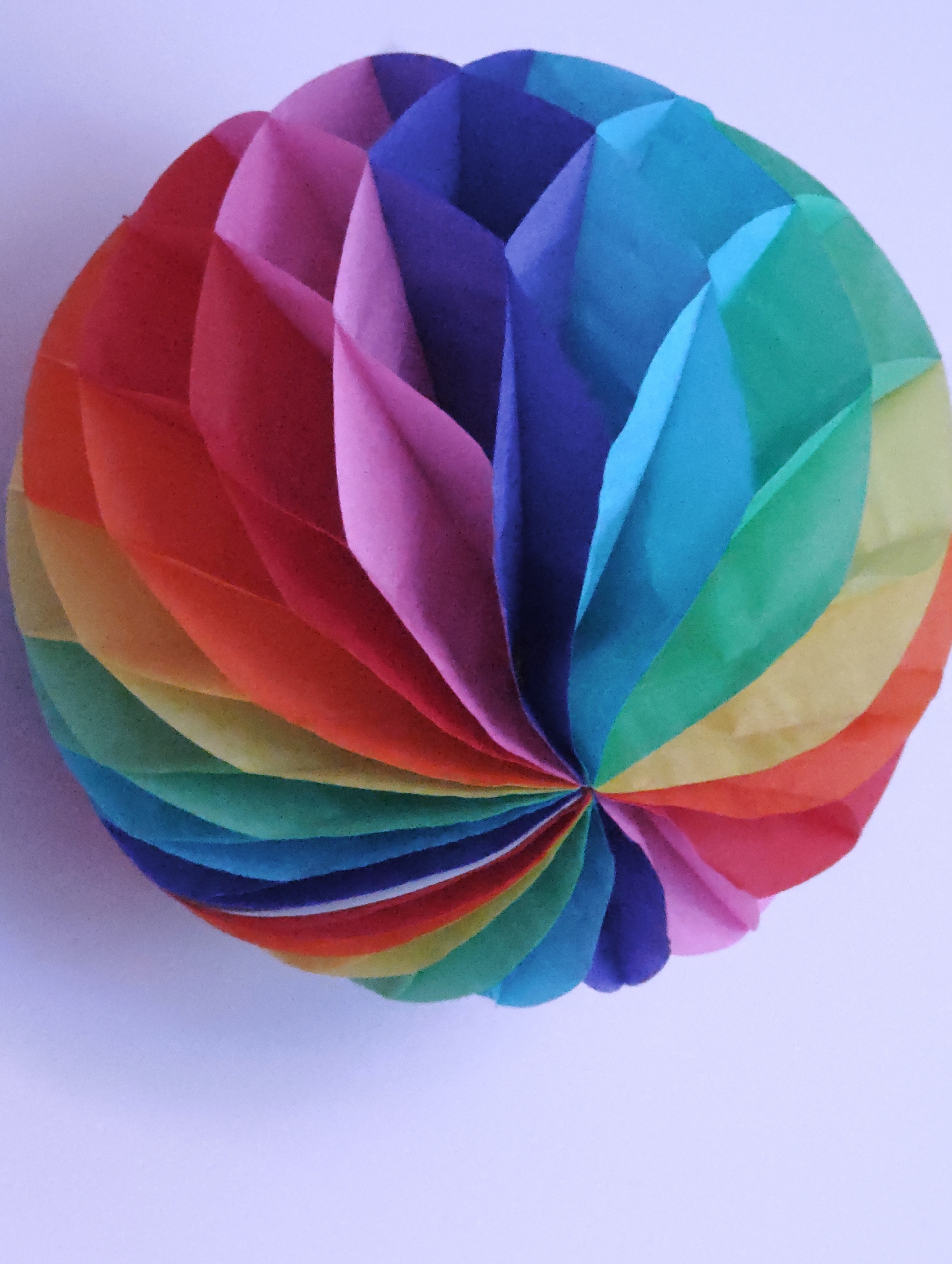 A circular, folded paper decoration with a rainbow of colors against a pale lavender background is shown. The decoration is composed of numerous pointed segments radiating from a central point, creating a fan-like appearance. The colors transition from red and orange on the left, through yellow, green, blue, and purple on the right. The background is a plain, smooth surface with a slight gradient of light. The image is a close-up shot, with the decoration filling most of the frame and casting a subtle shadow. The scene is static and does not contain any people. The location in Locking Parklands, United Kingdom, is not discernible from the visual information provided in the image.